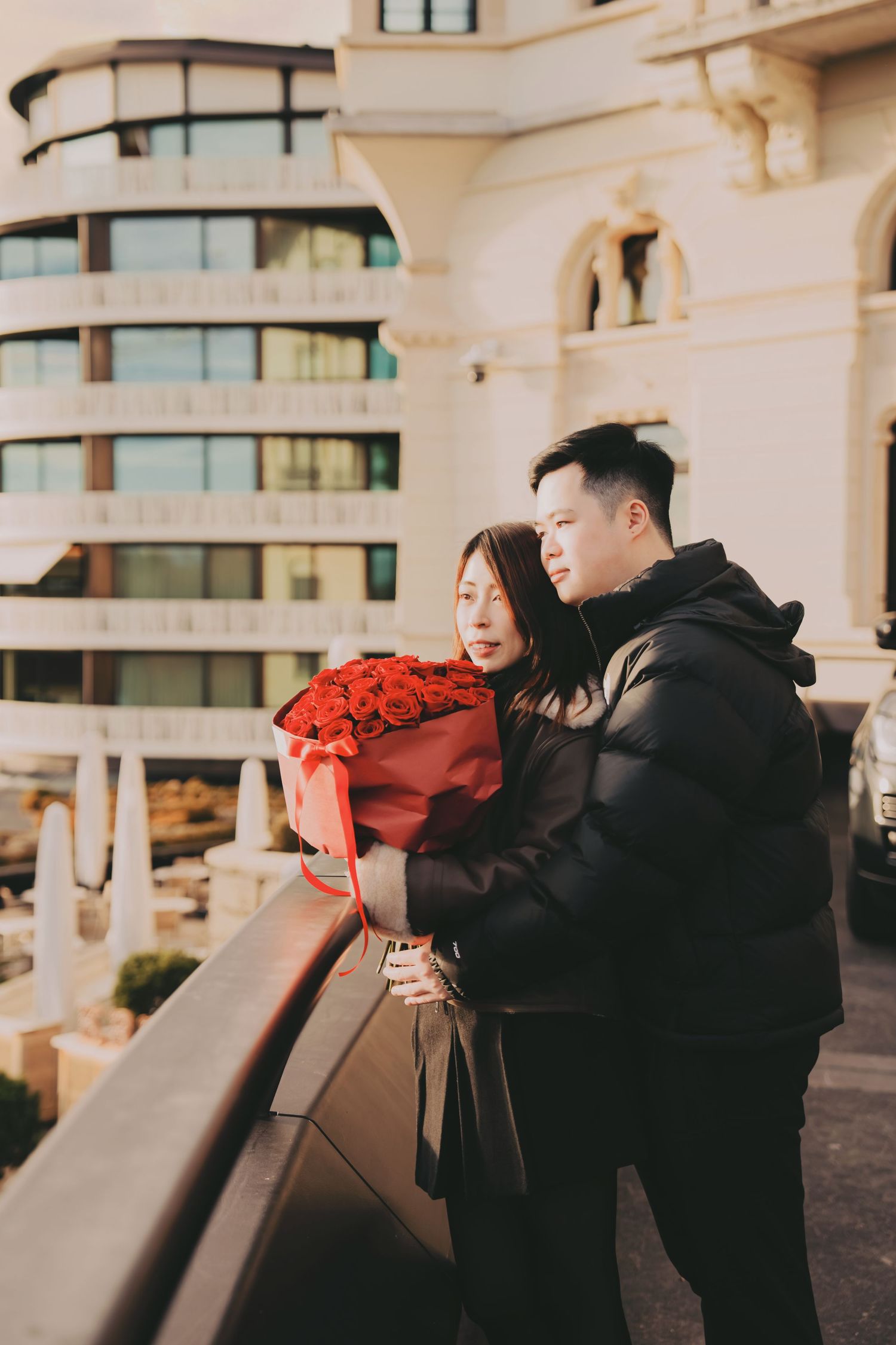 Asian couple sharing a romantic moment on a balcony with a large bouquet of red roses.