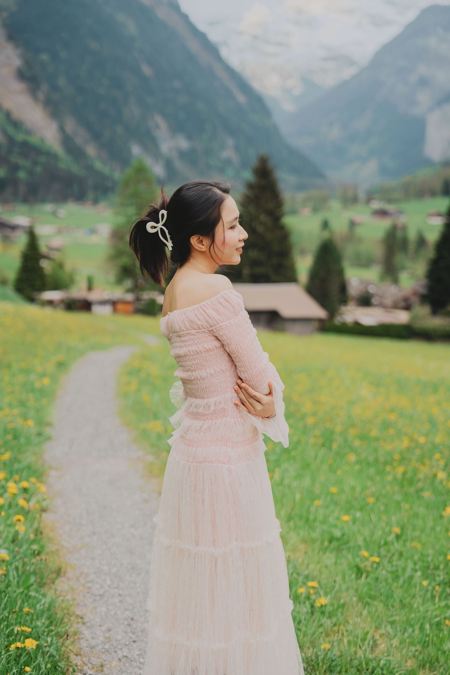 Woman in elegant pink off-shoulder dress standing in Swiss alpine meadow with mountain valley backdrop.