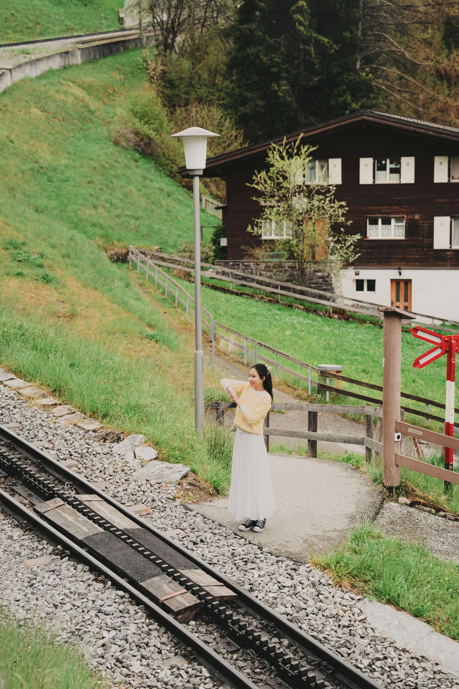 Woman in yellow top and white skirt standing near railway tracks beside a Swiss chalet and lamppost.