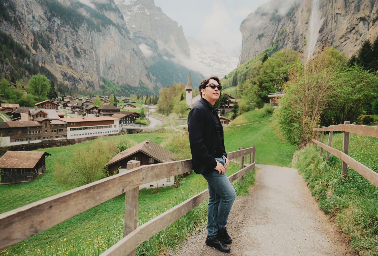 Man in black jacket and sunglasses standing on scenic path with picturesque Swiss alpine village in background.