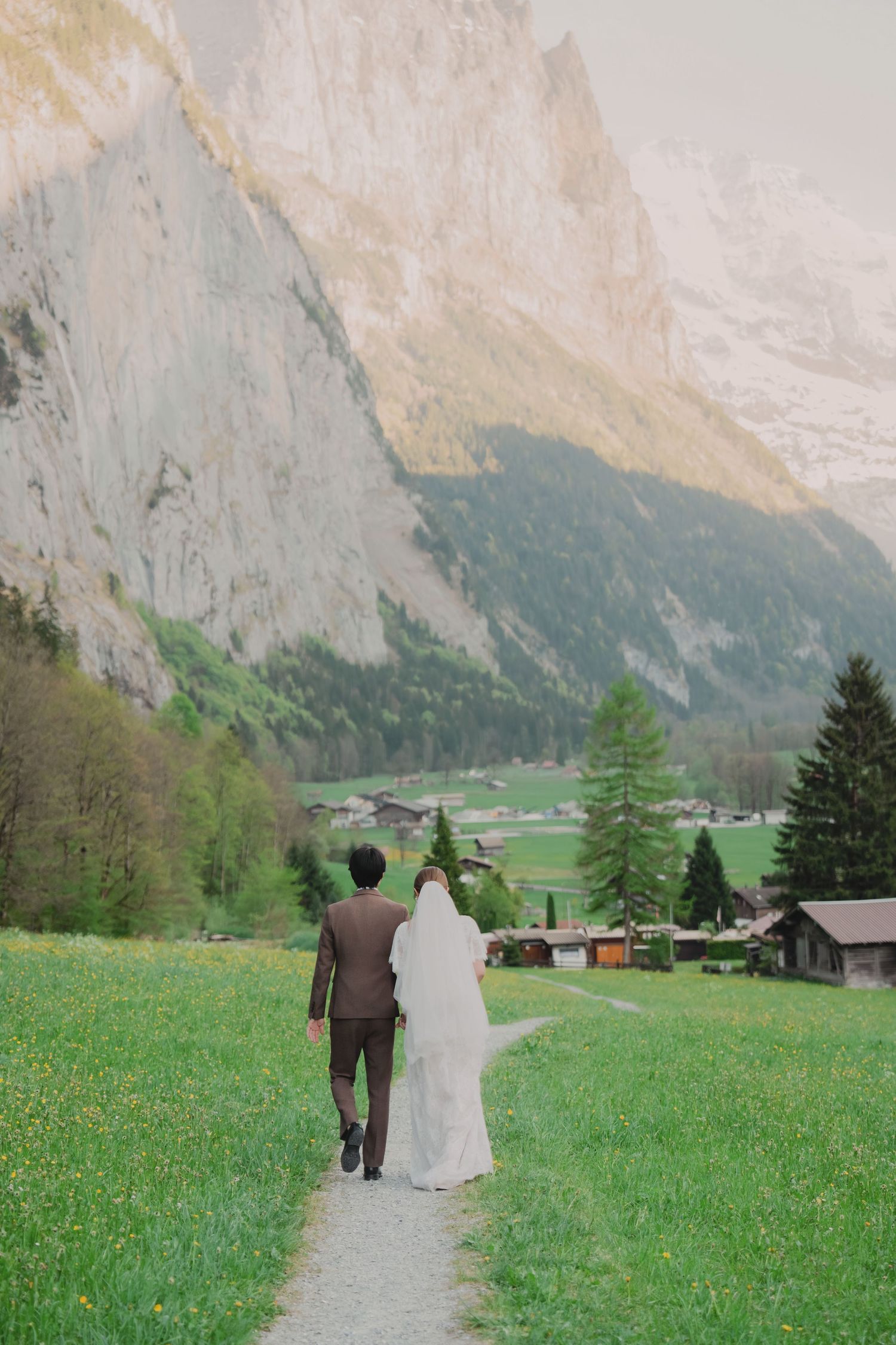 Bride and groom strolling together along a winding path through a lush Swiss alpine valley with towering cliffs.
