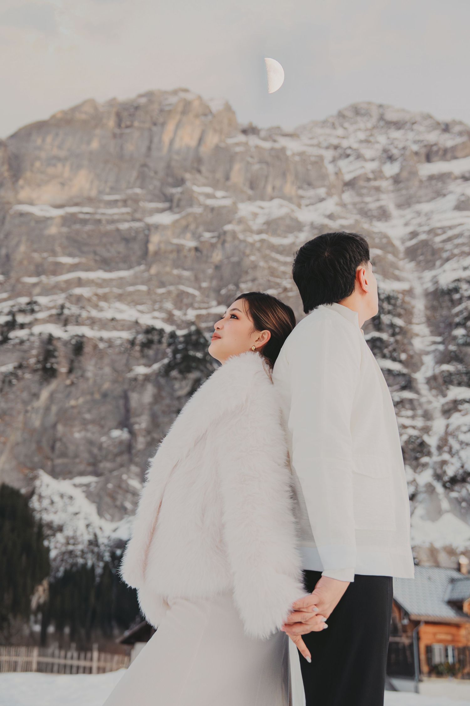 Couple holding hands back-to-back in white outfits with snowy mountain and crescent moon backdrop.