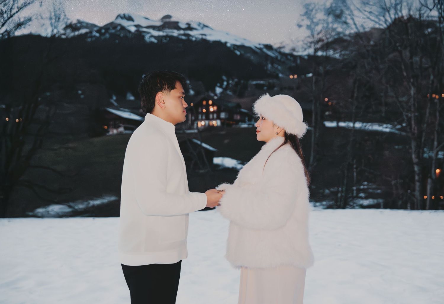 Couple holding hands in white winter outfits with snow-covered mountains at twilight.