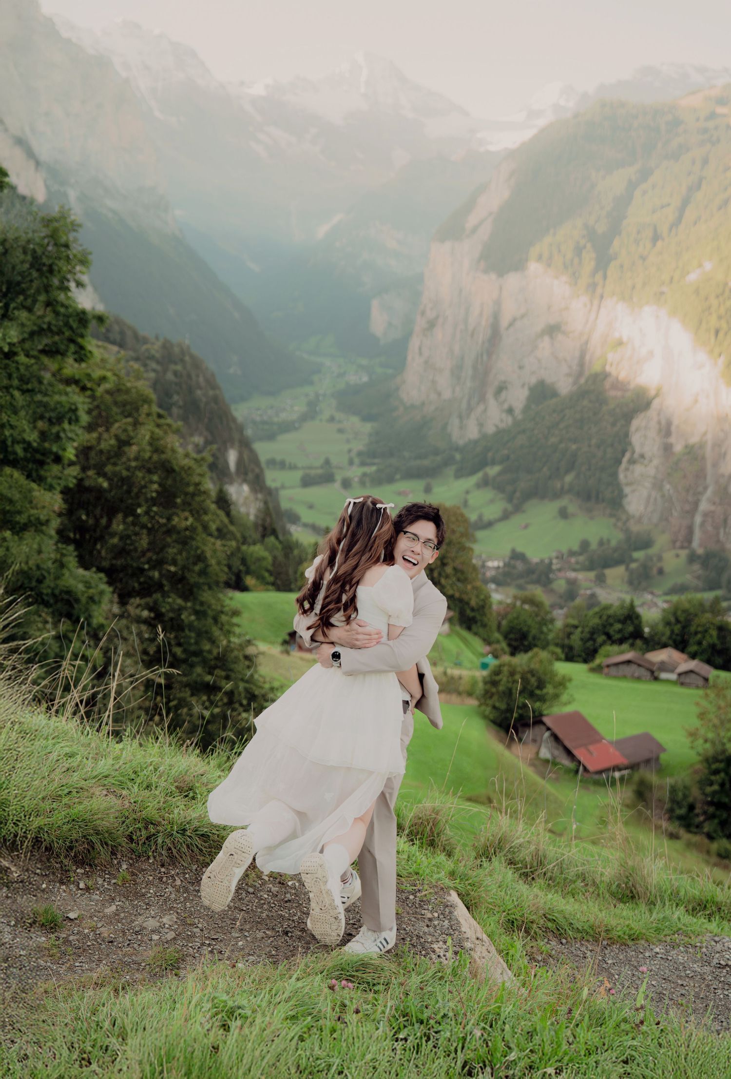 Bride being lifted joyfully on a Swiss Alpine hillside with a stunning green valley and mountain backdrop.