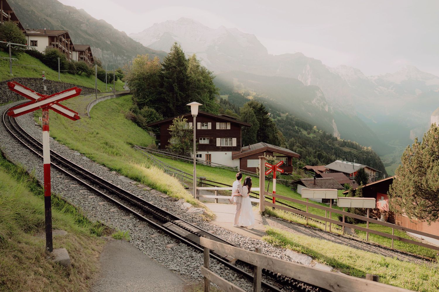 Bride in white gown stands beside a mountain railway track in a picturesque Swiss alpine village with misty peaks.