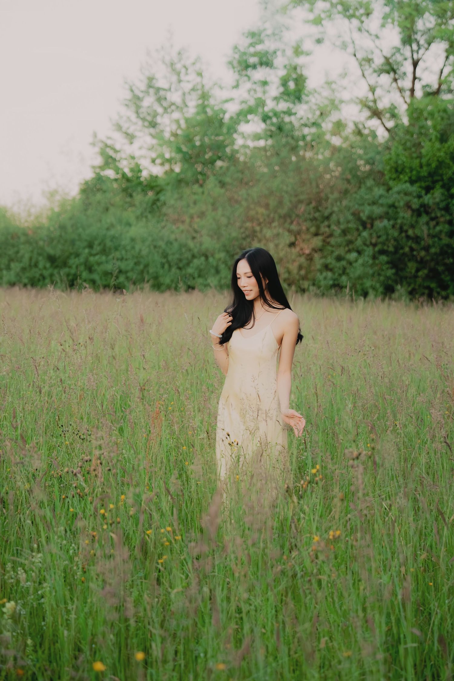 Woman in cream dress walking through tall green meadow with wildflowers at dusk.