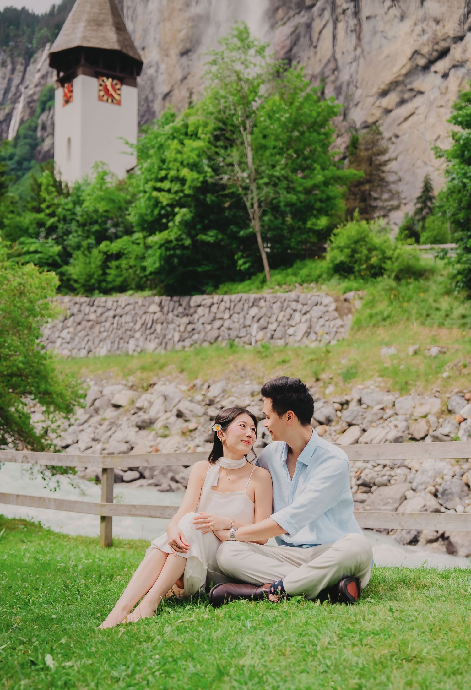 Couple sitting on grass by a scenic Swiss village with a historic clock tower and rocky cliffs.