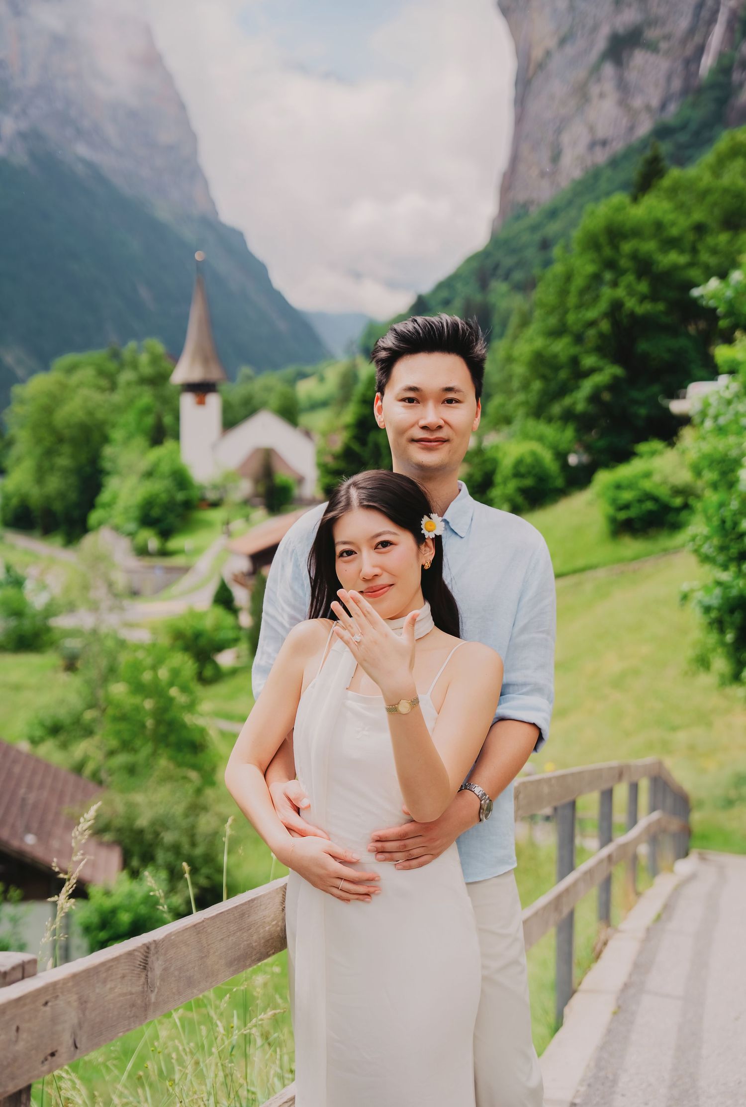 Couple posing on a wooden bridge with a picturesque Swiss village church and mountain backdrop.