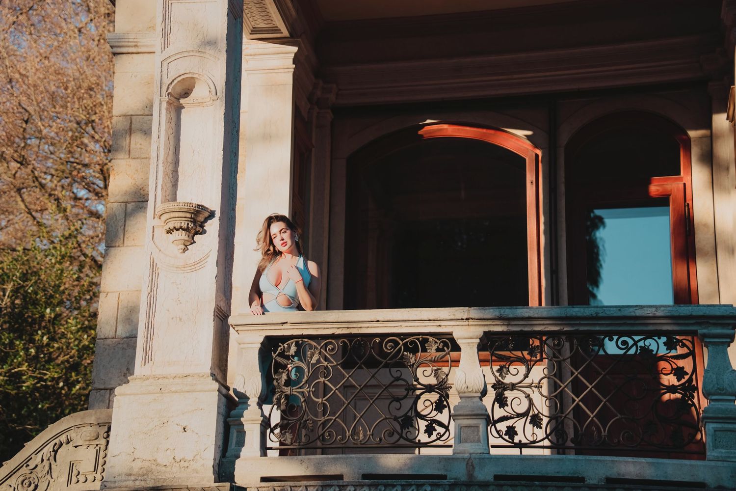 Woman in colorful dress stands on ornate iron balcony of a historic stone building in sunlight.