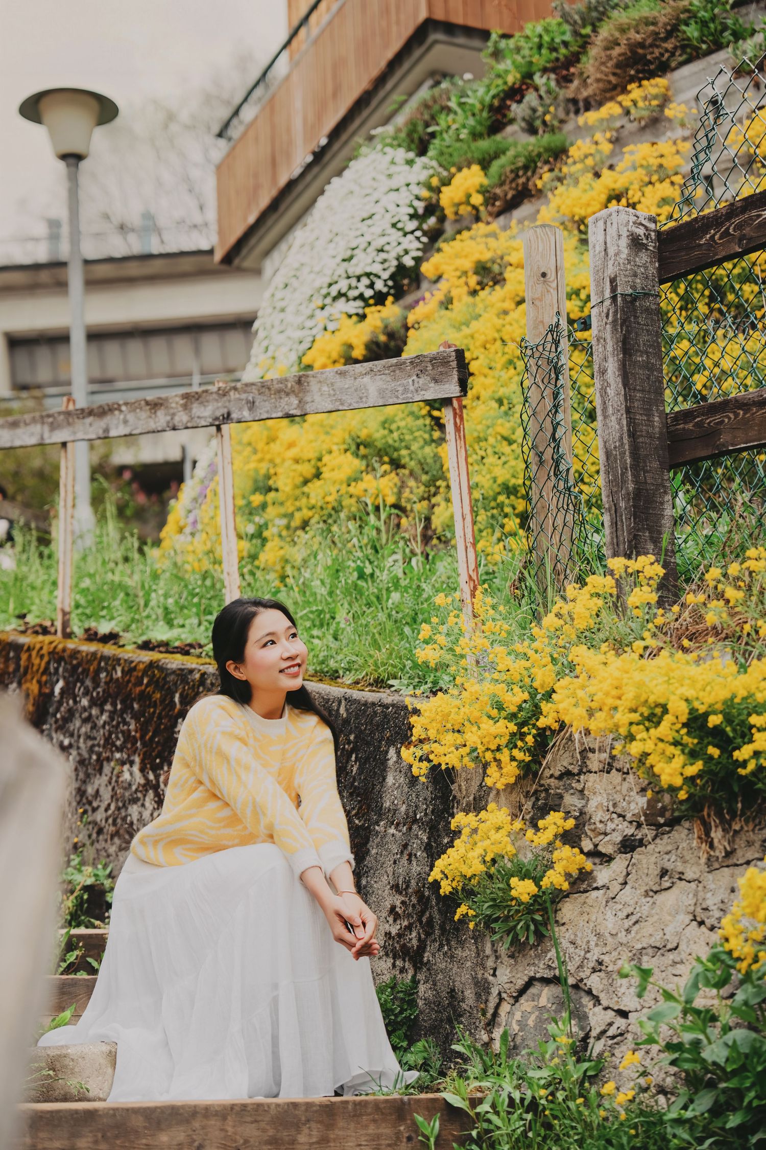 Young woman in yellow sweater and white skirt sitting by yellow wildflowers on a stone wall outdoors.