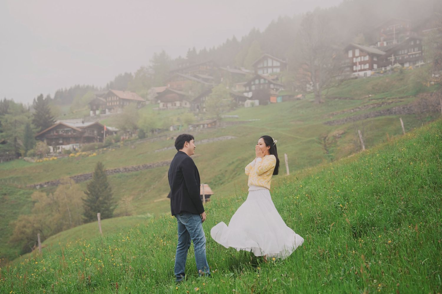 Couple sharing a romantic moment on a lush green hillside with misty Alpine village in background.