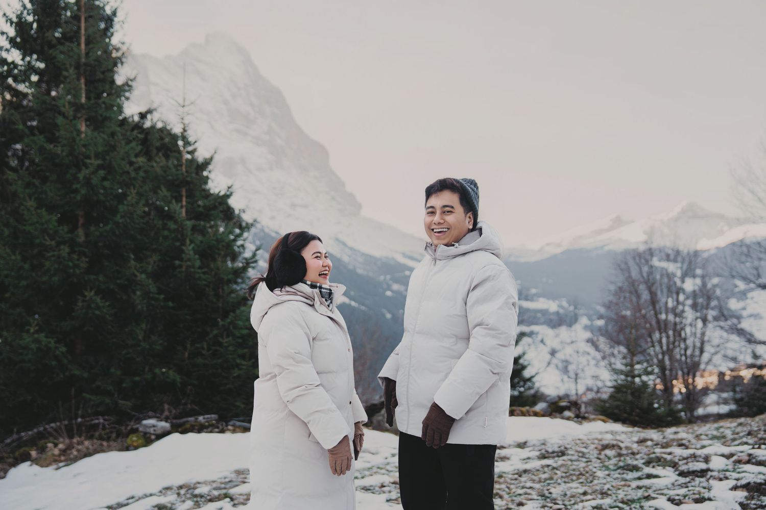 Smiling couple in white puffer jackets holding hands with snowy mountain peak visible behind them.