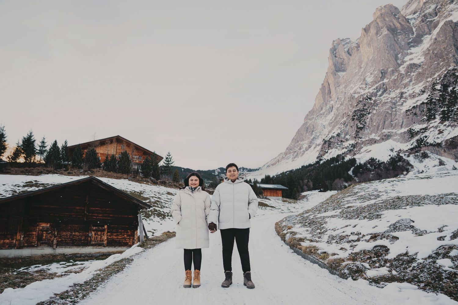 Couple standing hand in hand on snowy alpine path with wooden chalet and rocky mountain backdrop.