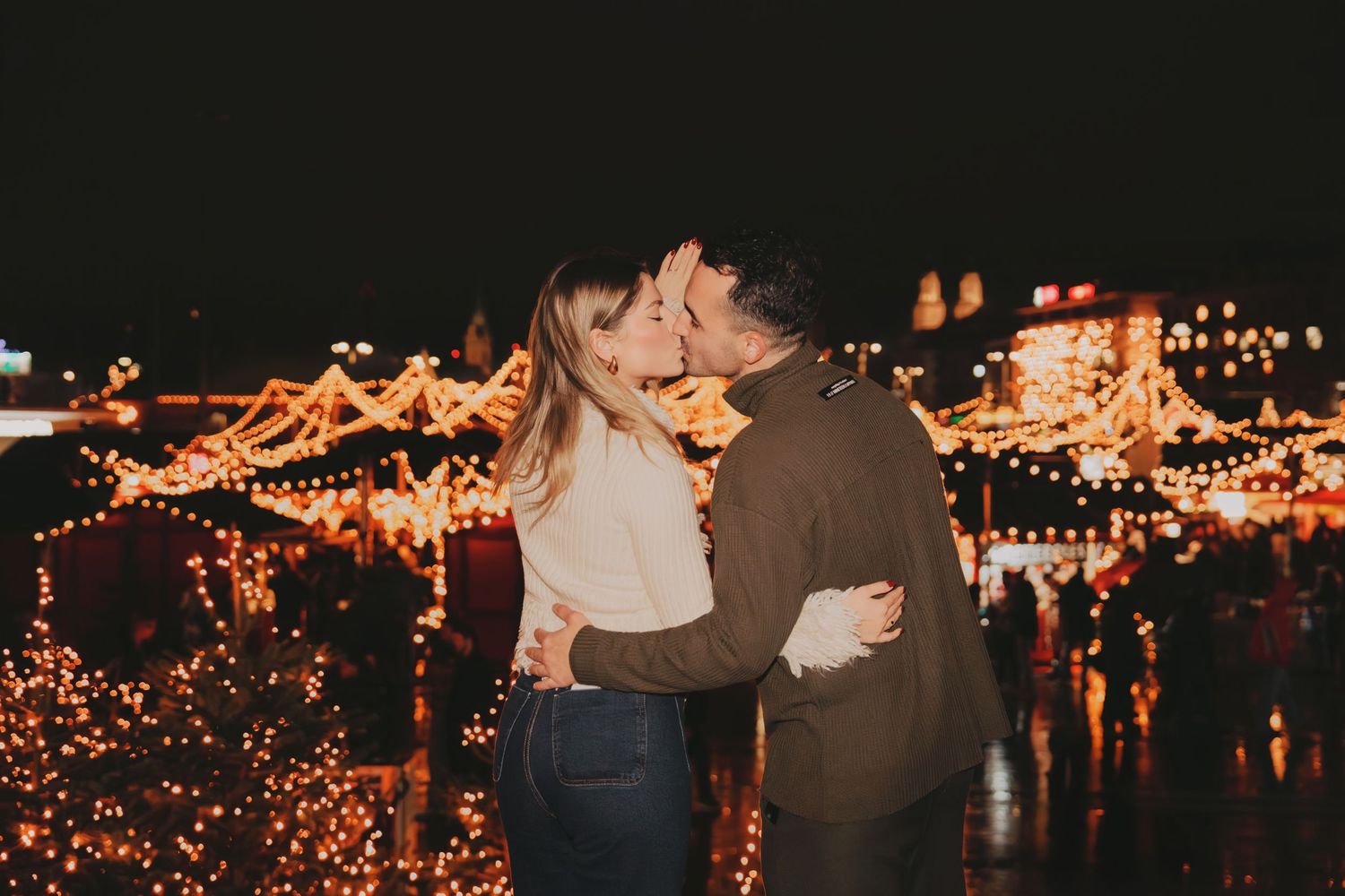 Couple sharing a kiss at a romantic Christmas market glowing with warm festive lights at night.