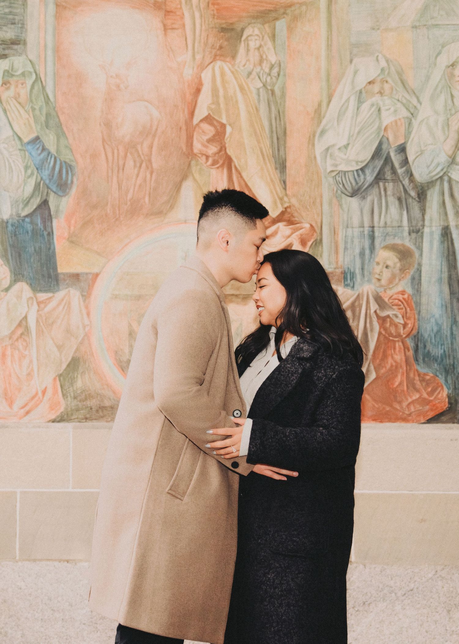 Newly engaged couple embracing and kissing in front of a pastel-toned religious fresco.