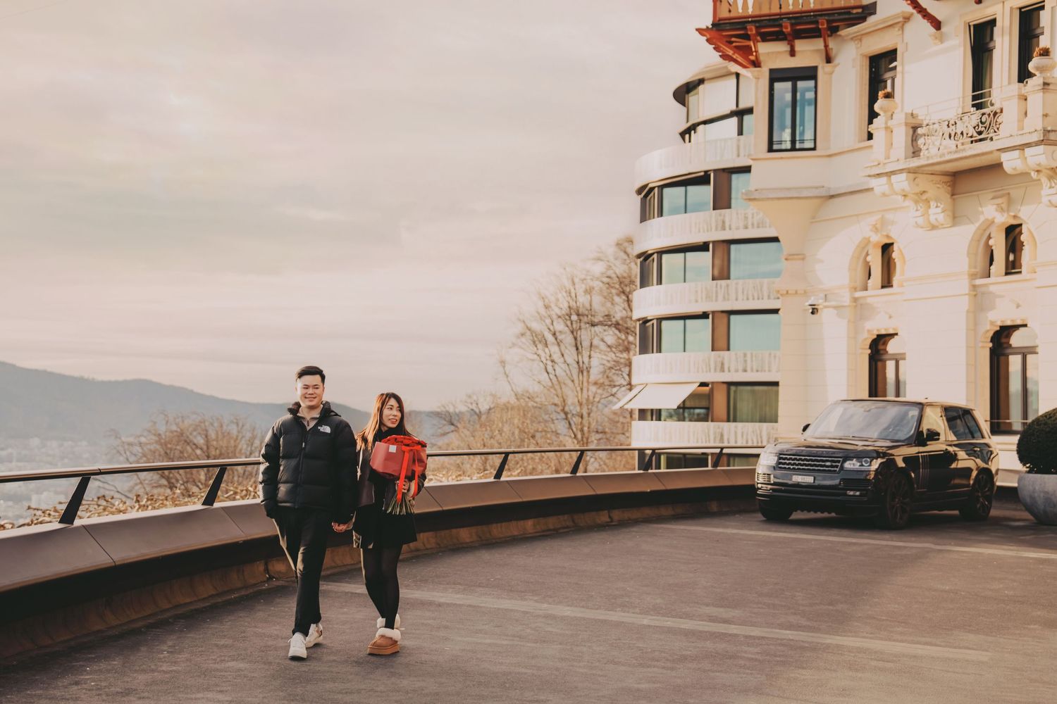 Couple posing confidently on an elevated terrace beside a luxury SUV with scenic mountain backdrop.