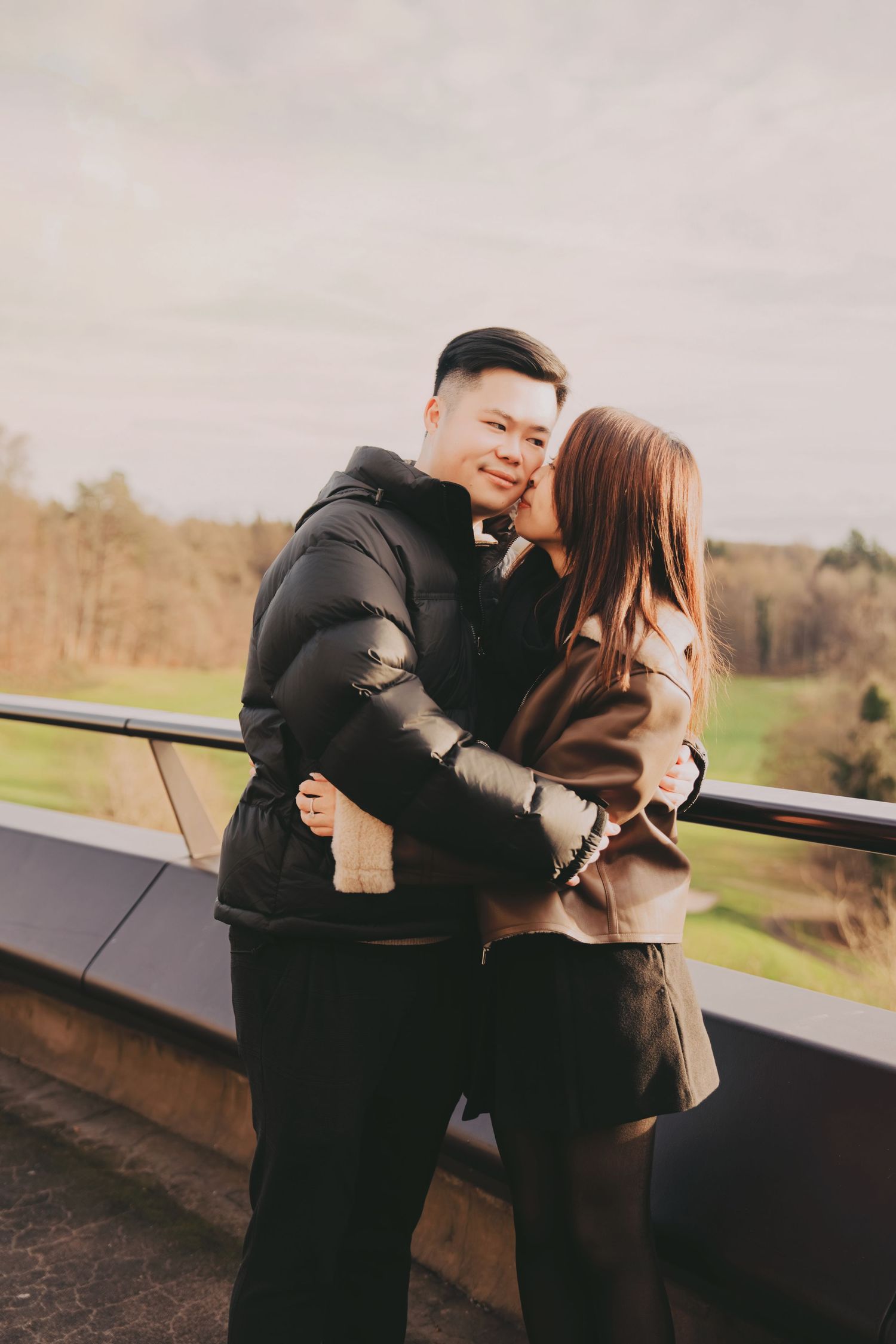 Young Asian couple embracing on balcony with scenic green landscape behind them.