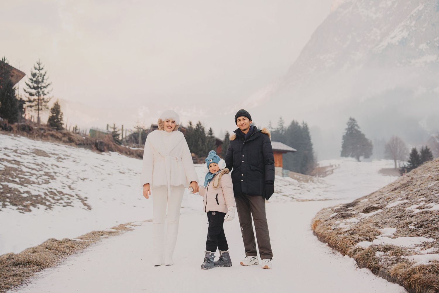 Family of three holding hands on snowy mountain road with pine trees and misty peaks in background.