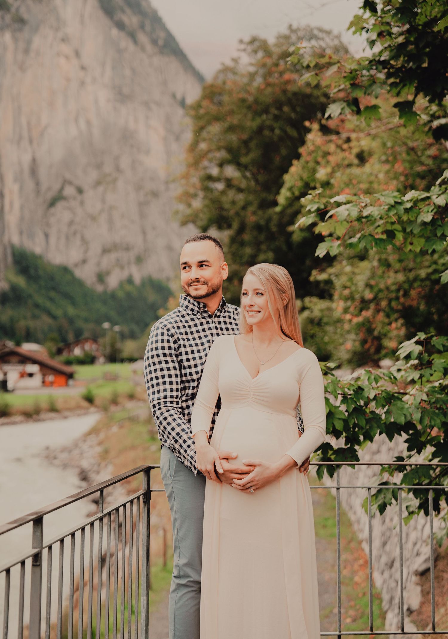 Happy couple standing together on bridge, pregnant woman in cream dress with Swiss mountains behind.