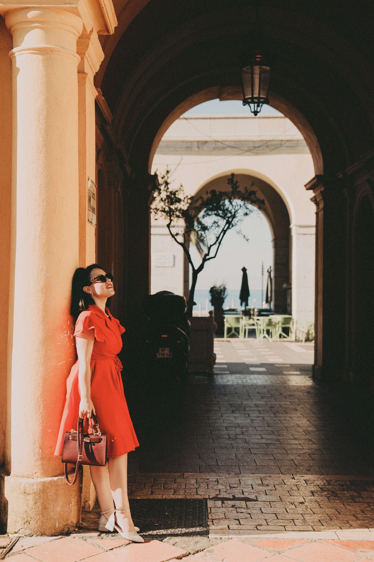 Stylish woman in red dress with handbag posing under ornate arches in Monaco arcade.