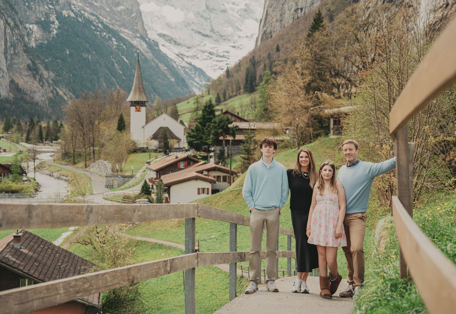 Family portrait on a wooden walkway overlooking a charming Swiss alpine village with mountains and a church steeple.