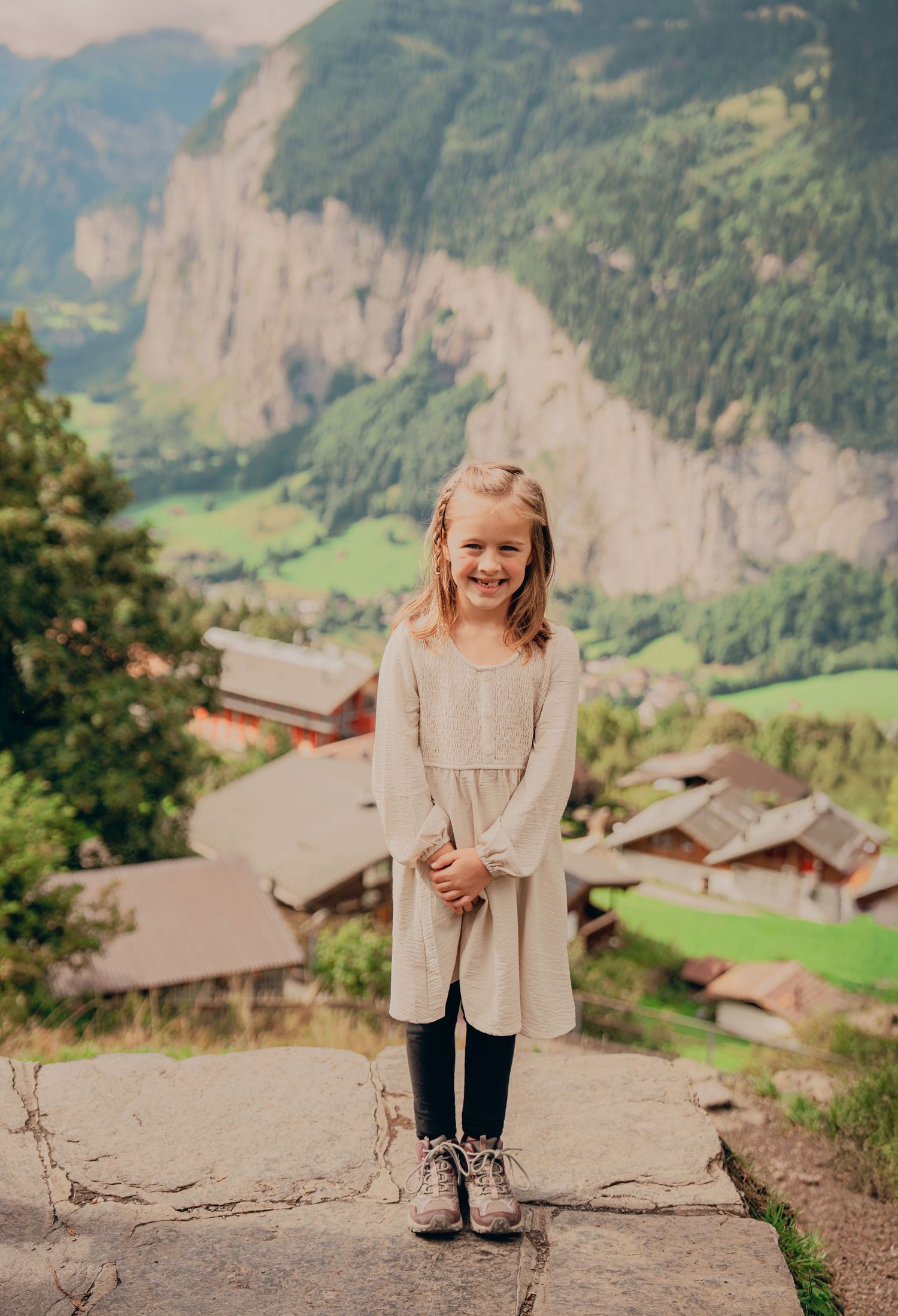 Cheerful girl in beige dress and boots smiling on stone wall with Swiss Alpine village in background.