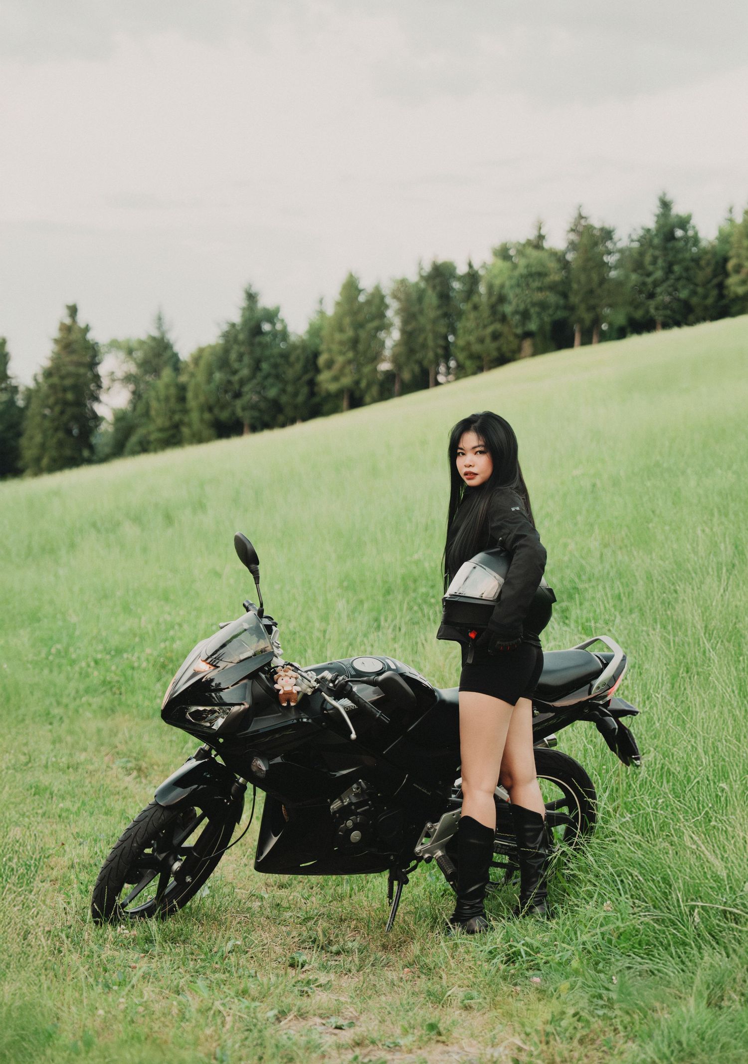 Young woman in black outfit holding helmet beside black sport motorcycle on green hillside.
