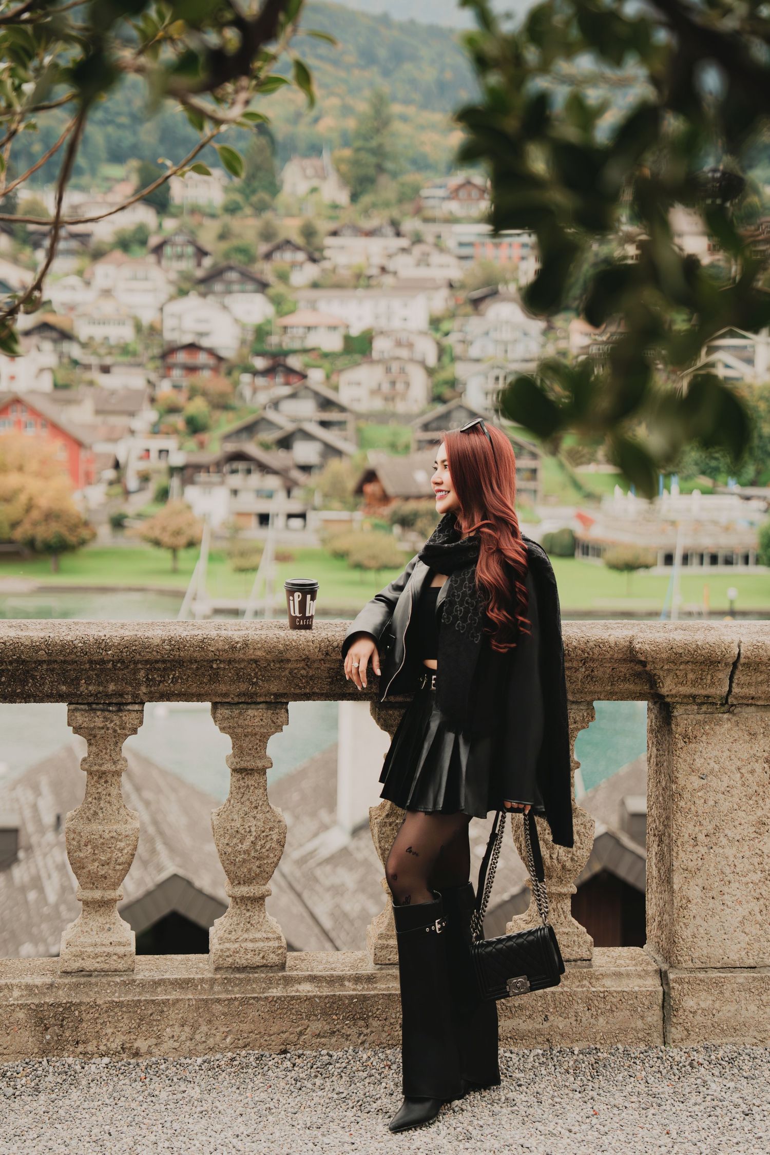 Fashionable woman in black leather skirt and cape holds coffee at stone balustrade above Swiss village.