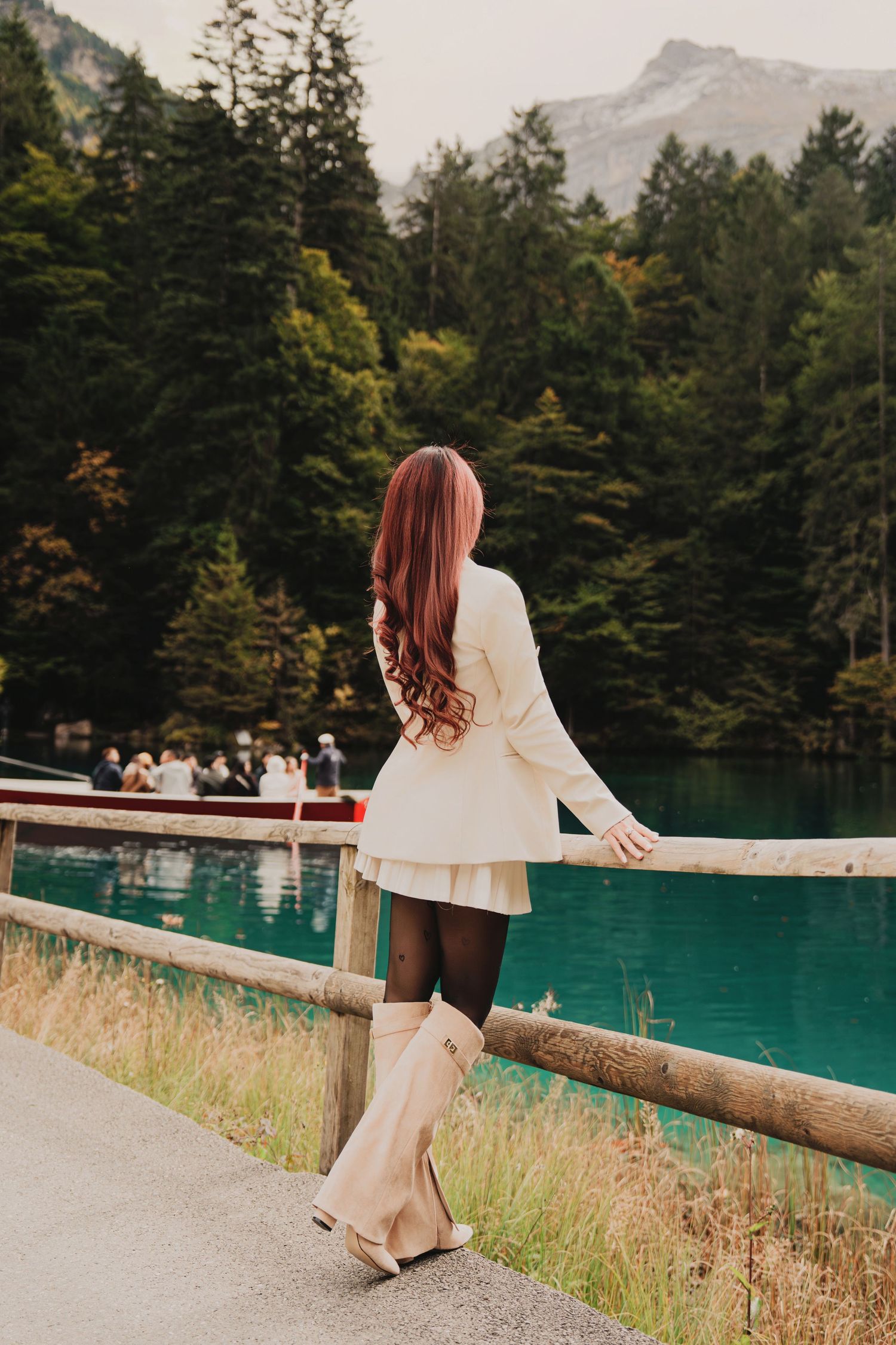 Stylish woman with long red hair overlooking vivid turquoise lake framed by mountain forest.
