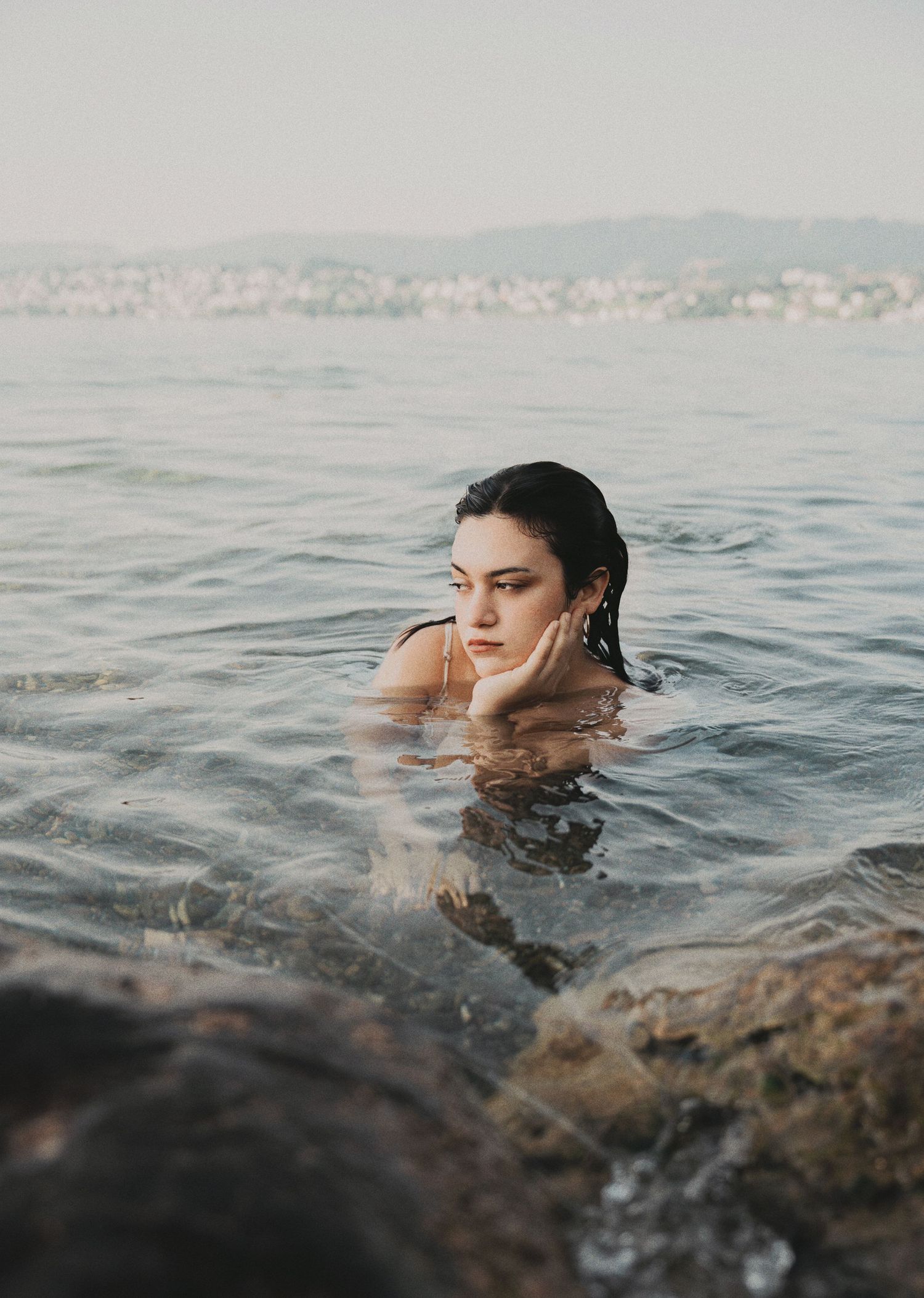 Young woman with wet dark hair resting chin on hand while swimming in calm sea water near rocky shore.