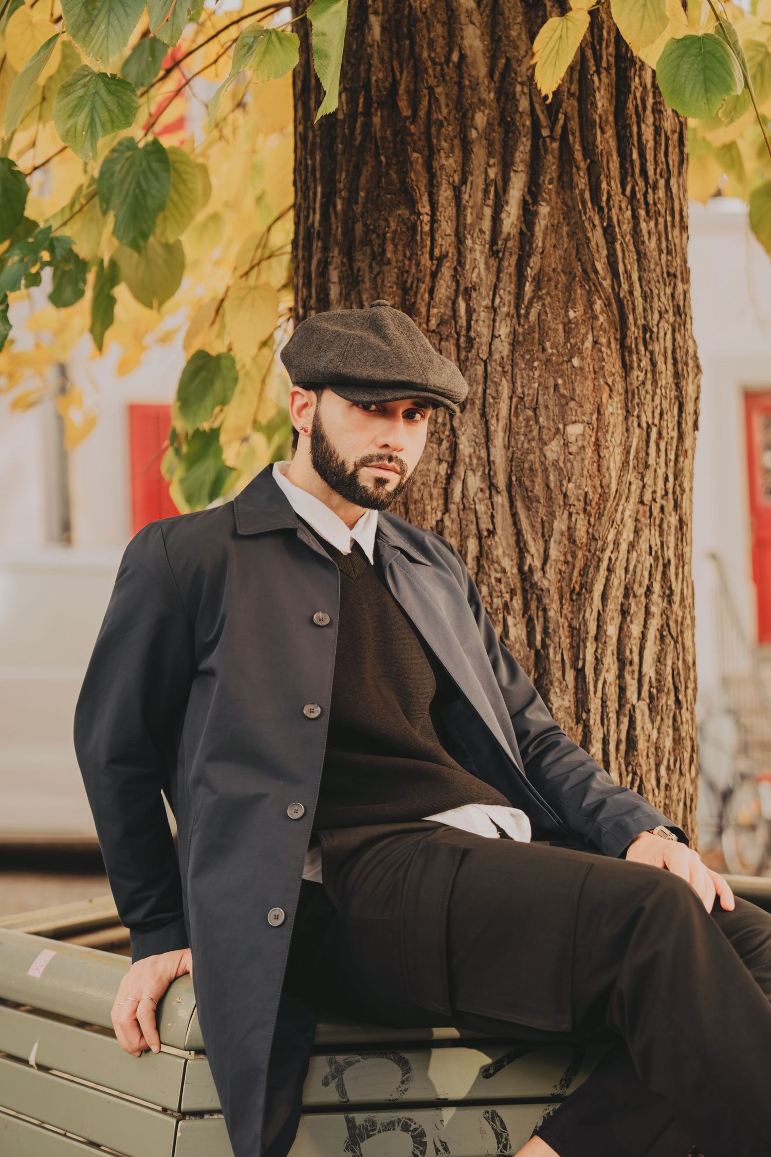 Bearded man in dark coat and flat cap sitting relaxed on bench beneath large tree in autumn park.