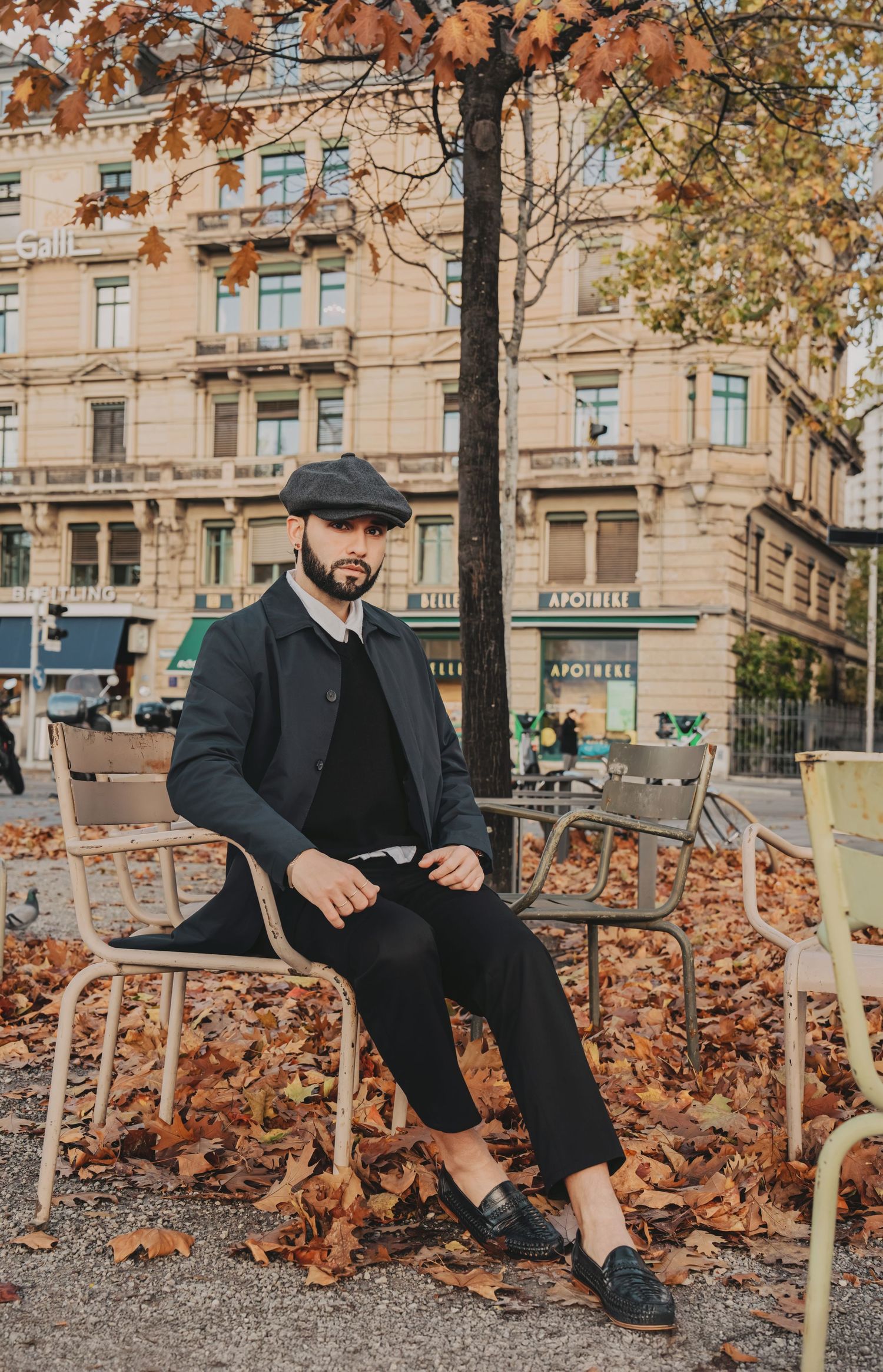 Trendy man in newsboy cap and dark coat sitting among fallen autumn leaves in city square.