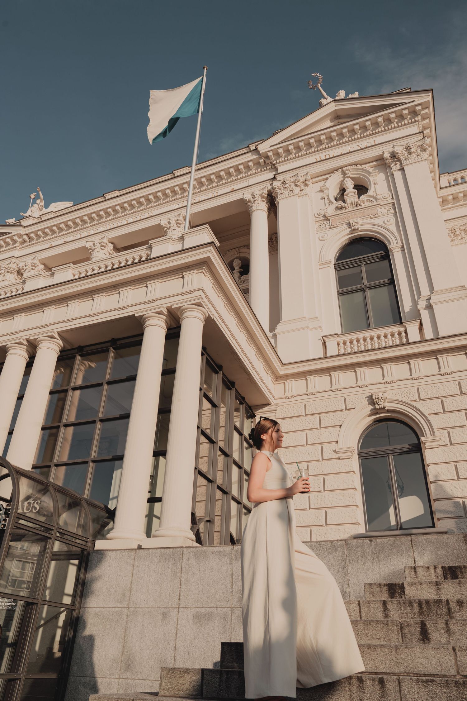 Woman in white dress standing on steps of neoclassical building with columns and flag.