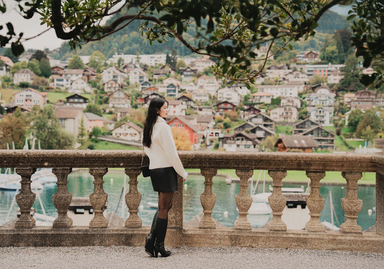 Woman in white sweater and black skirt gazes at picturesque Swiss lakeside village from stone balustrade.
