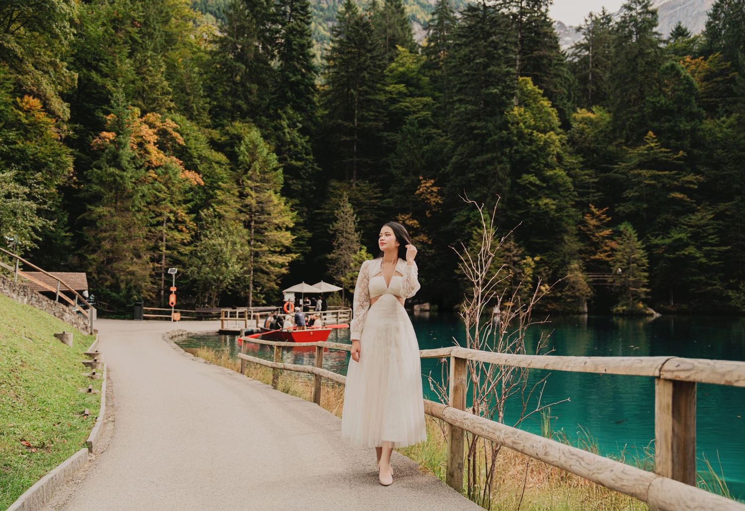 Young woman in cream dress walking along a path beside a stunning turquoise lake with forest scenery.