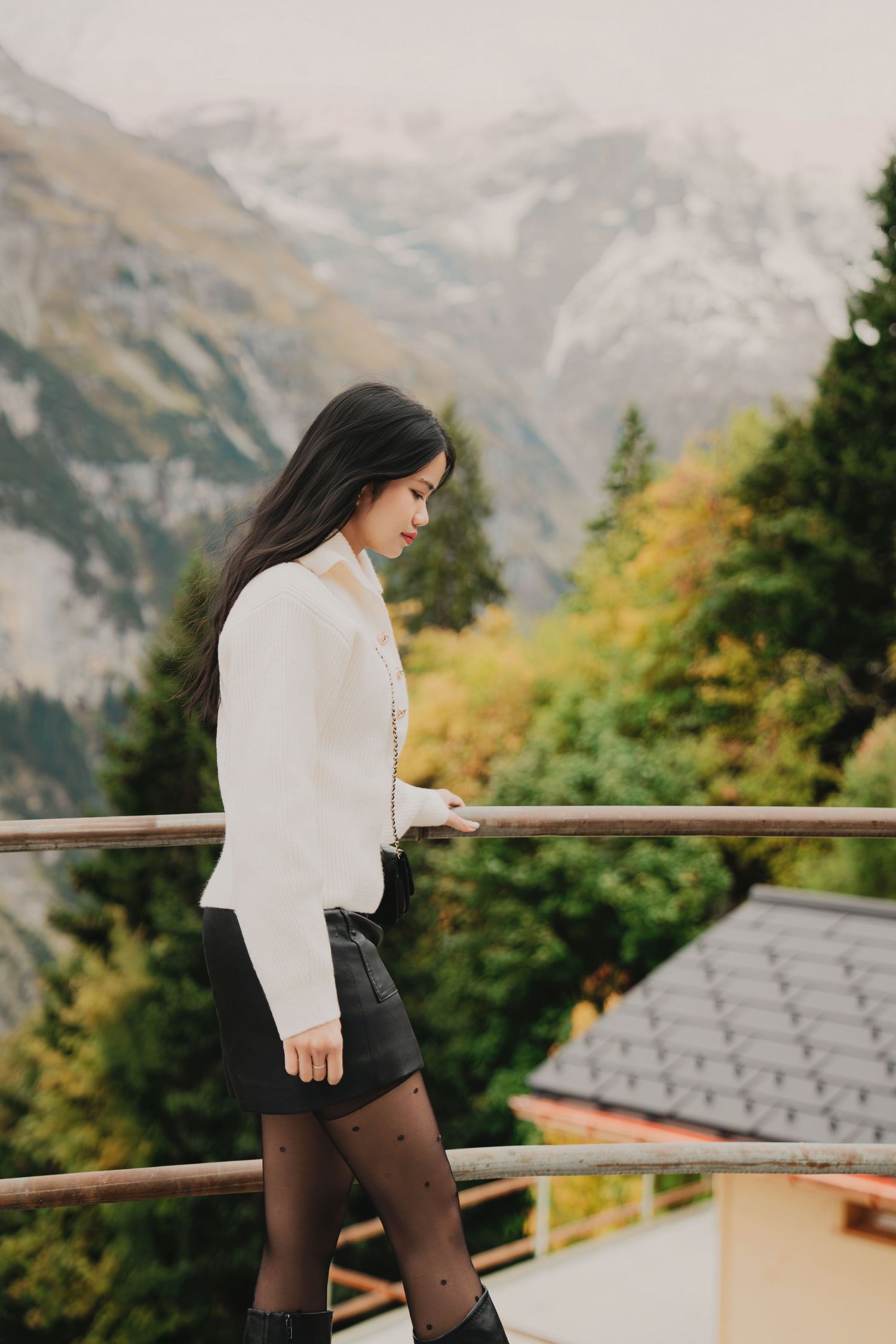 Young woman in white sweater and black shorts standing on wooden balcony with misty alpine mountains.