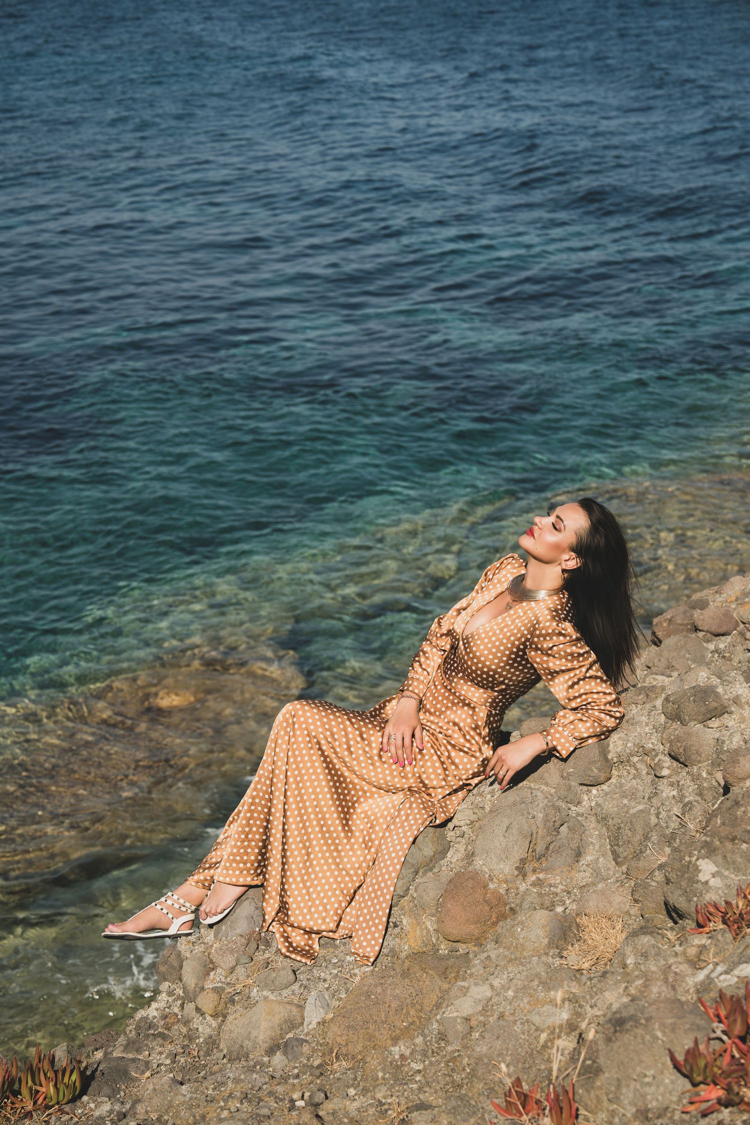 Elegant woman in gold polka dot dress reclining on coastal rocks with clear blue sea behind.