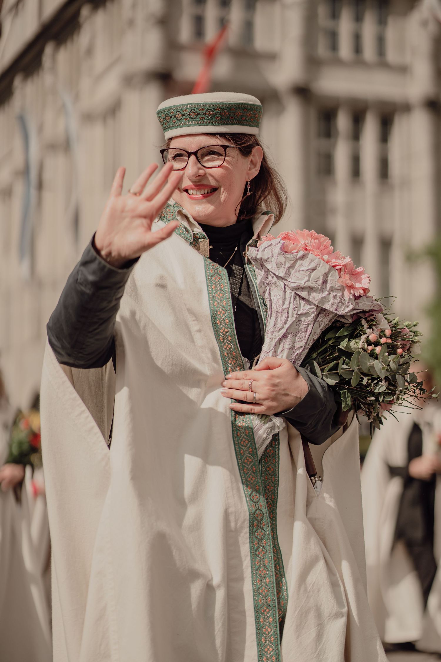 Smiling woman in white ceremonial robe with green-trimmed hat waves while holding flower bouquet.