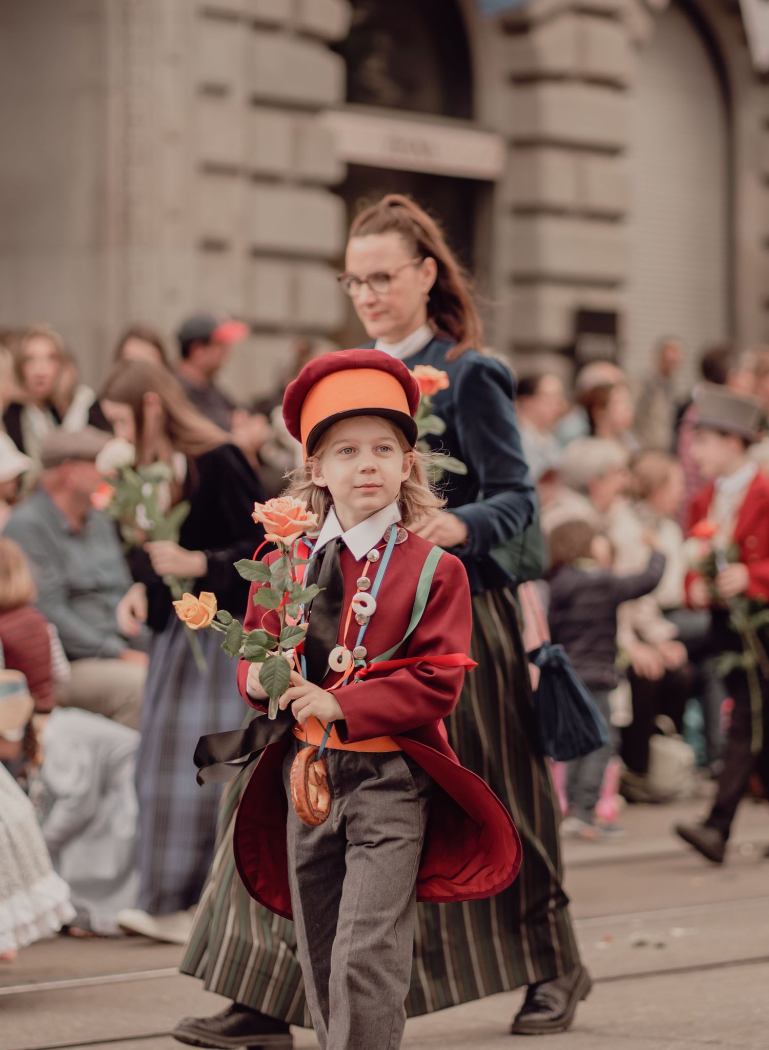 Young child in red vintage costume carries orange roses while marching in a traditional street parade.
