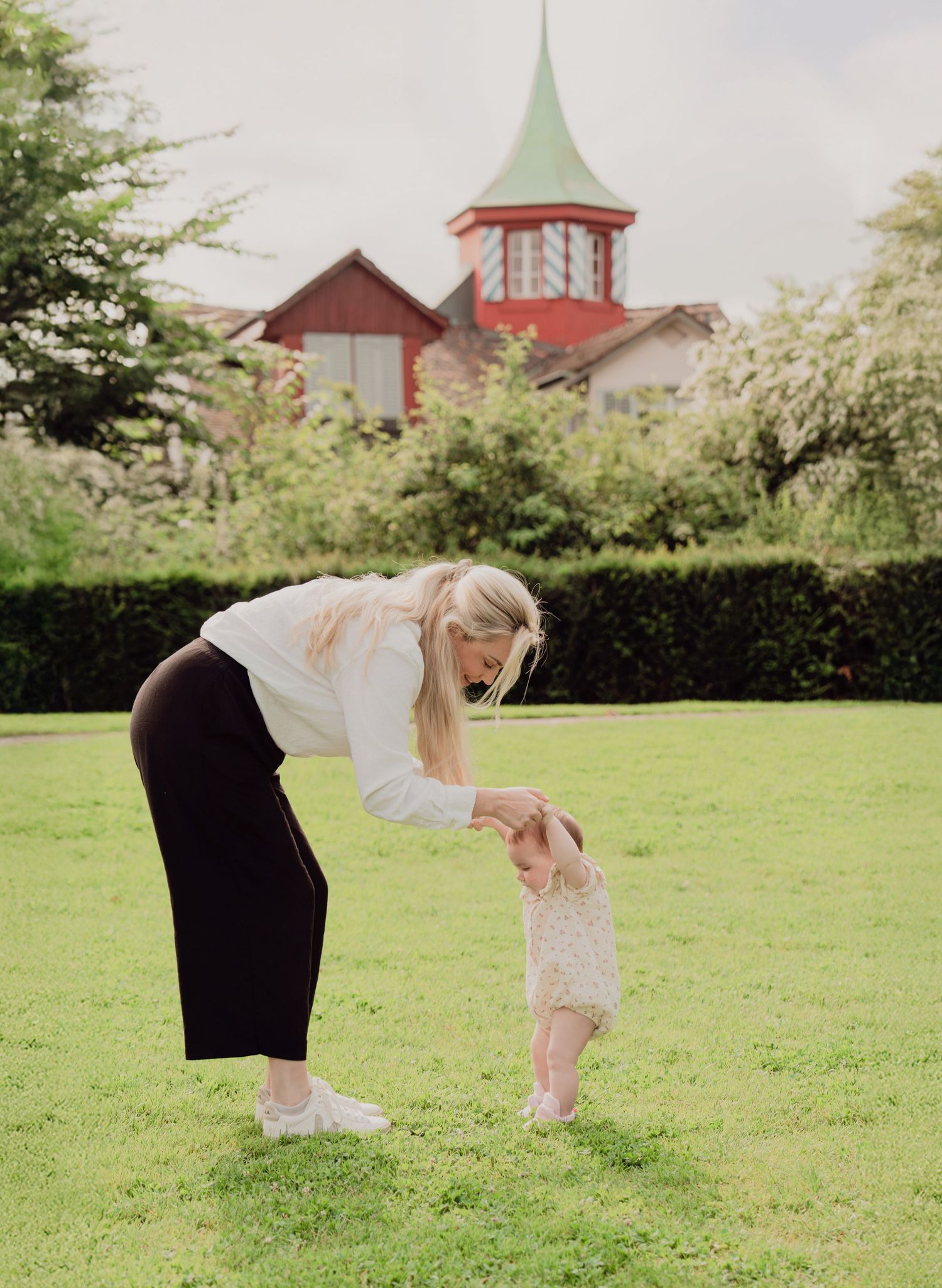 Blonde mother helping baby take first steps on green lawn with red-roofed building in background.