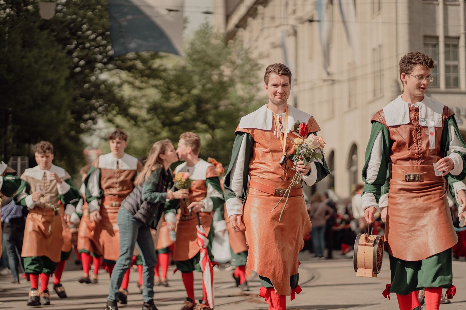 Young men in matching orange medieval costumes march in a traditional historical street parade carrying flowers.