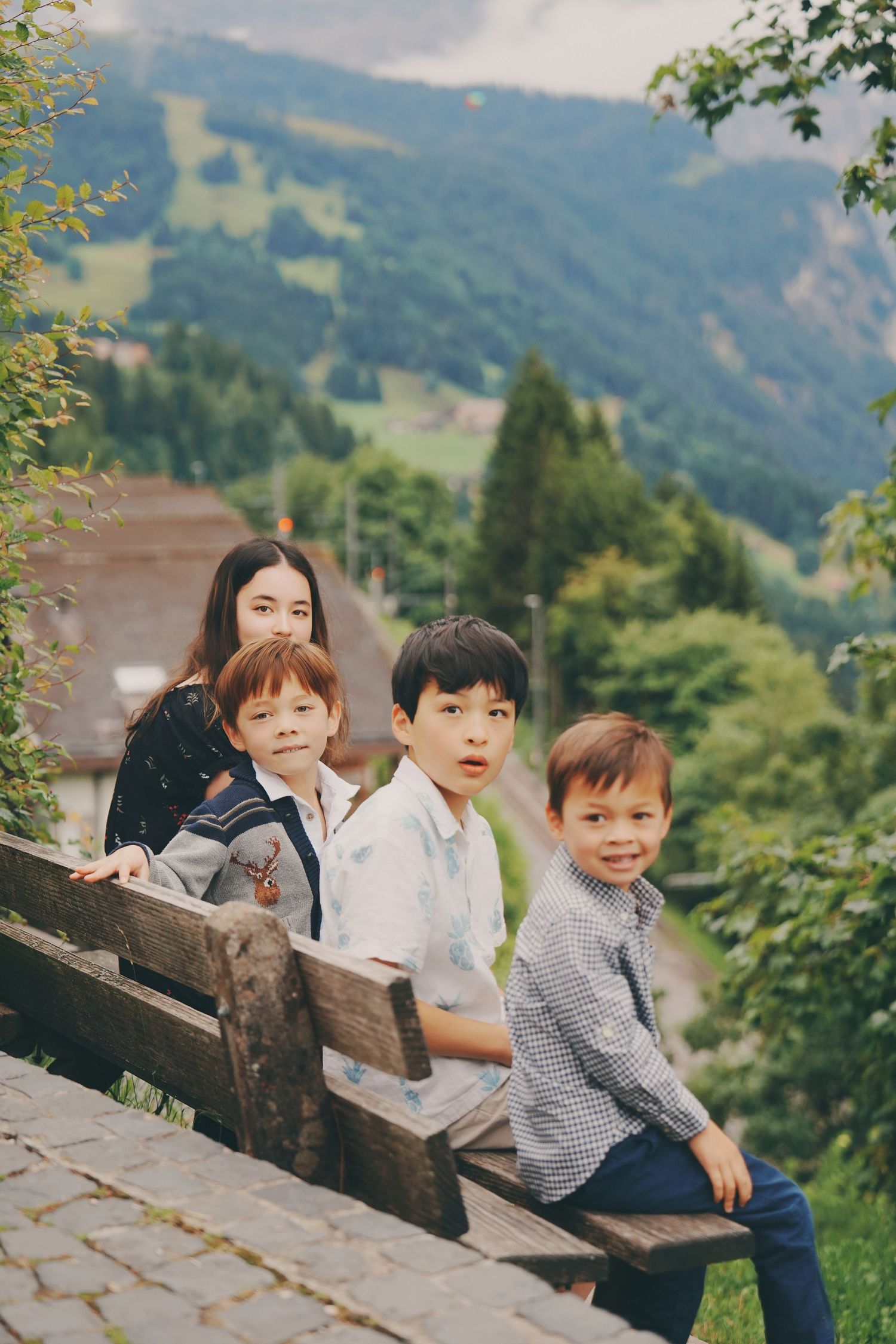 Four children pose on a wooden bench with scenic Swiss Alpine mountains and green valleys in the background.