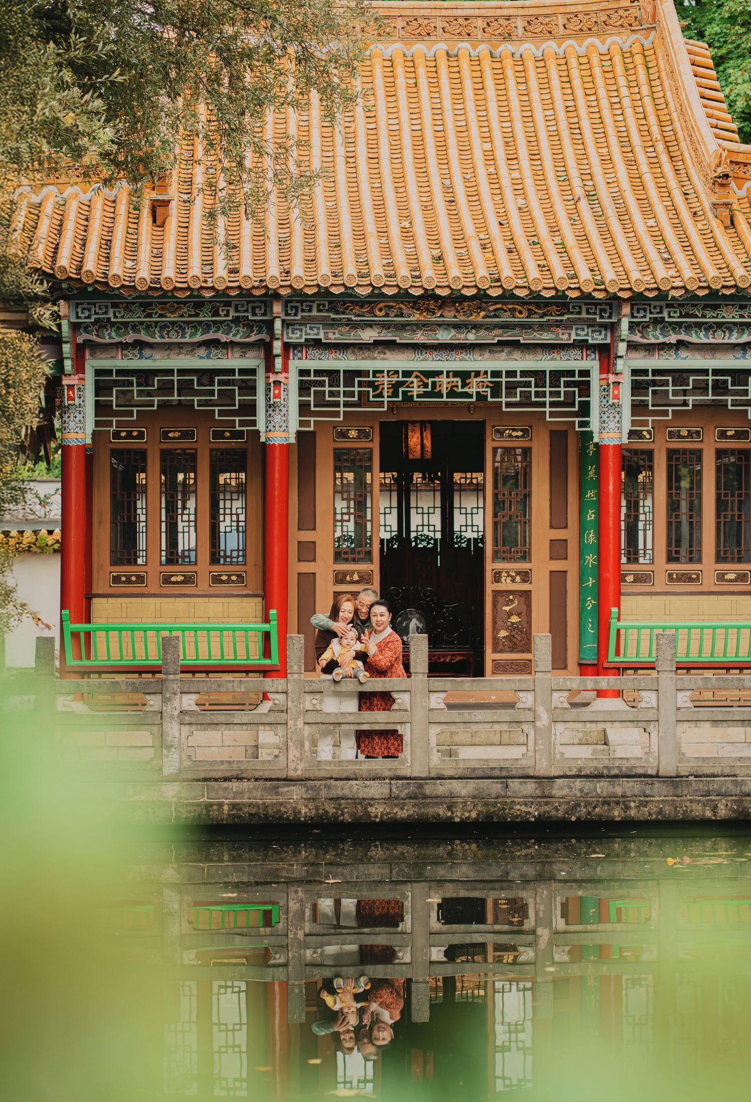 Family with baby reflected in pond before traditional Chinese pagoda with golden tiled roof.