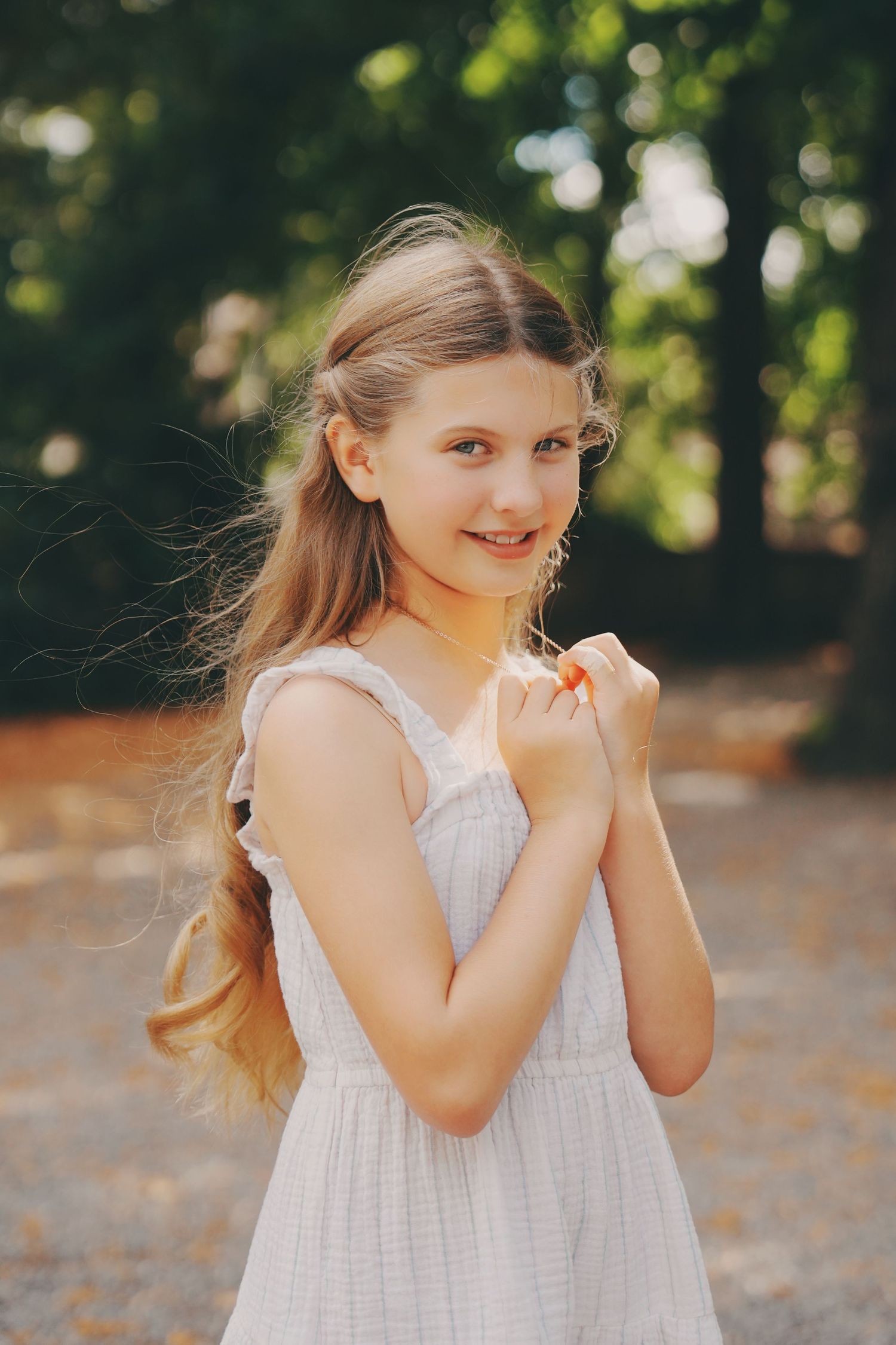 A cheerful young girl in a white dress stands in a sunny park with trees in the background.