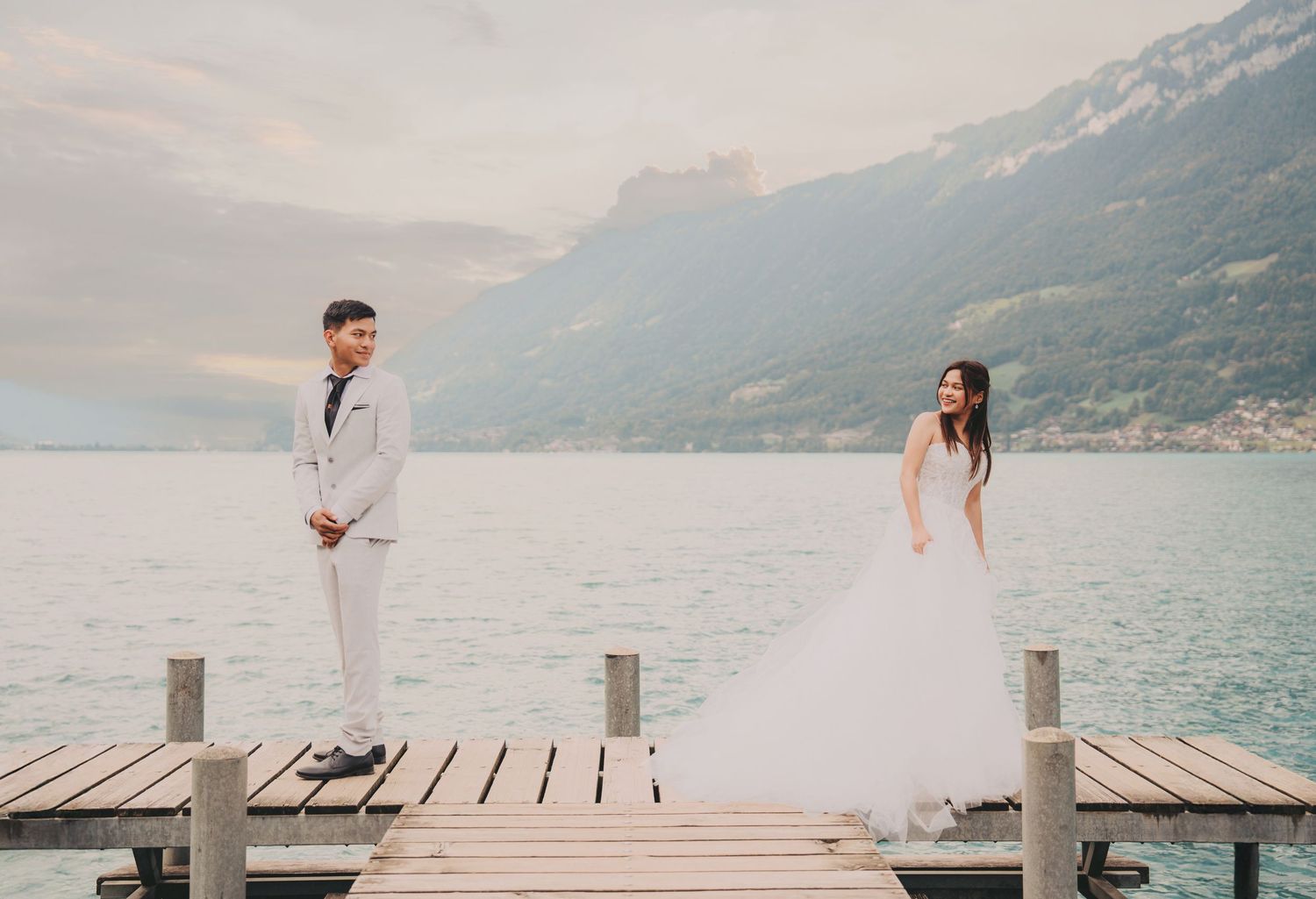 Elegant couple in wedding attire facing each other on a pier with scenic alpine lake views.