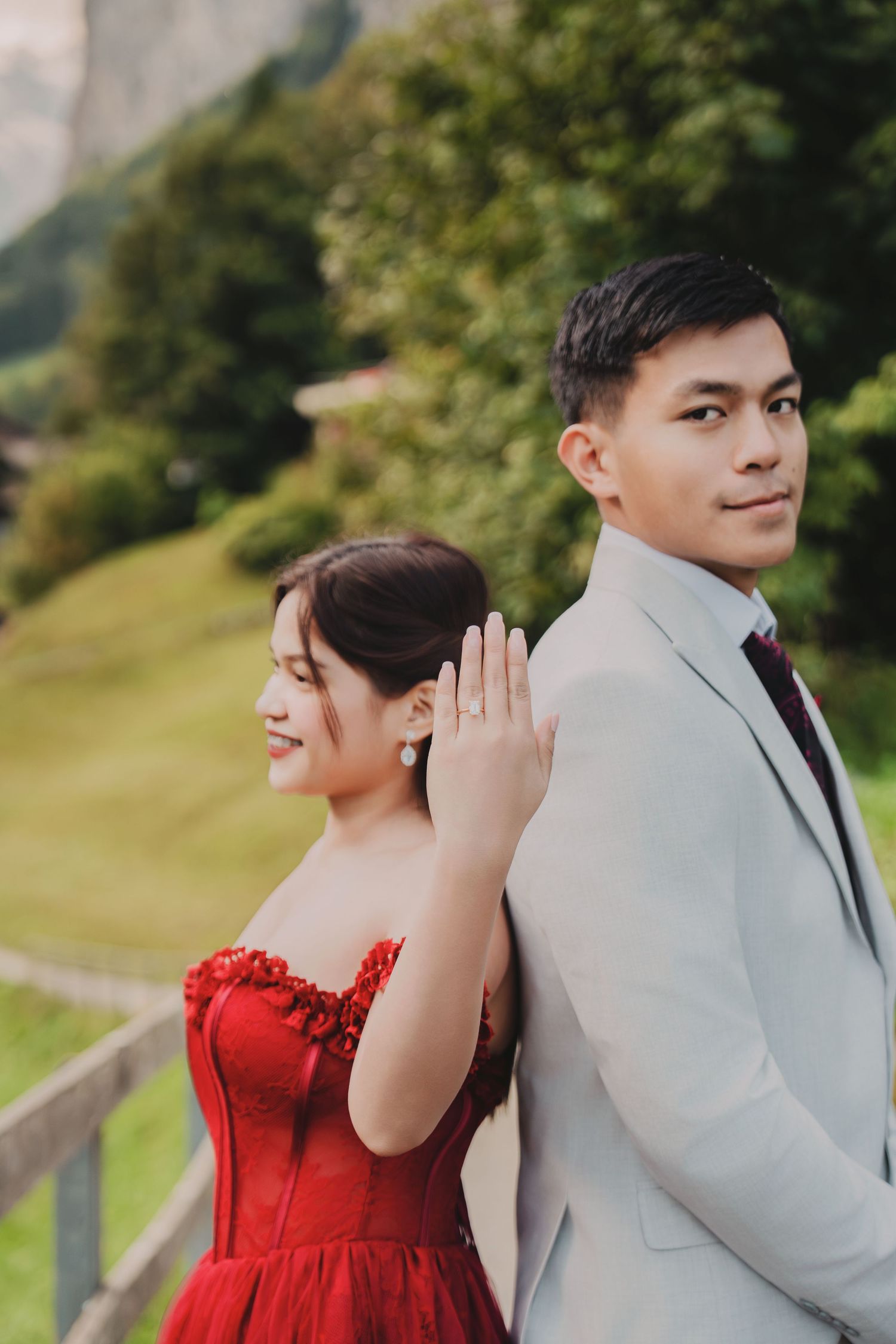 Couple standing back-to-back, bride showing engagement ring, with lush green Swiss mountain scenery.