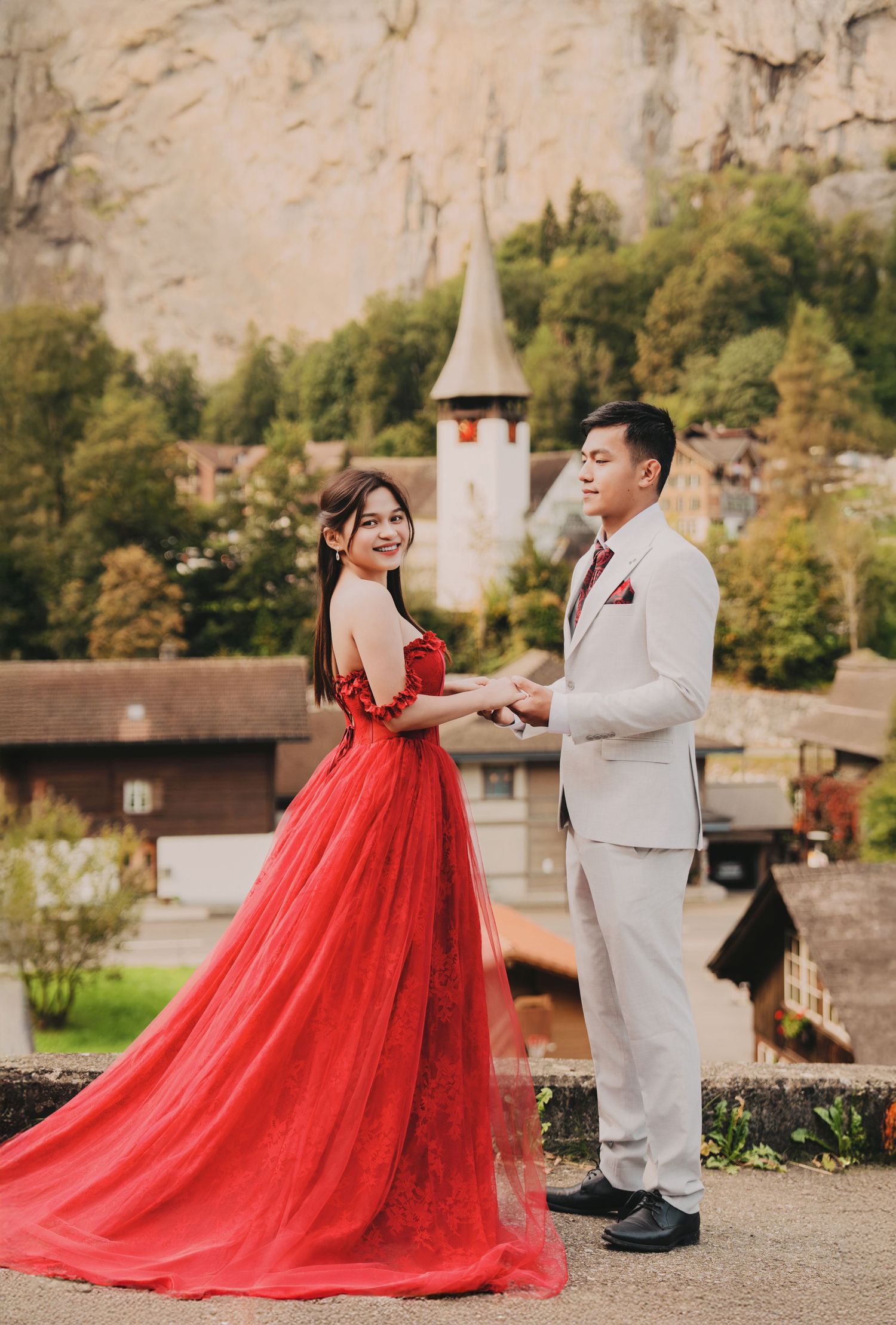 Couple in red gown and grey suit holding hands with Swiss village church backdrop.
