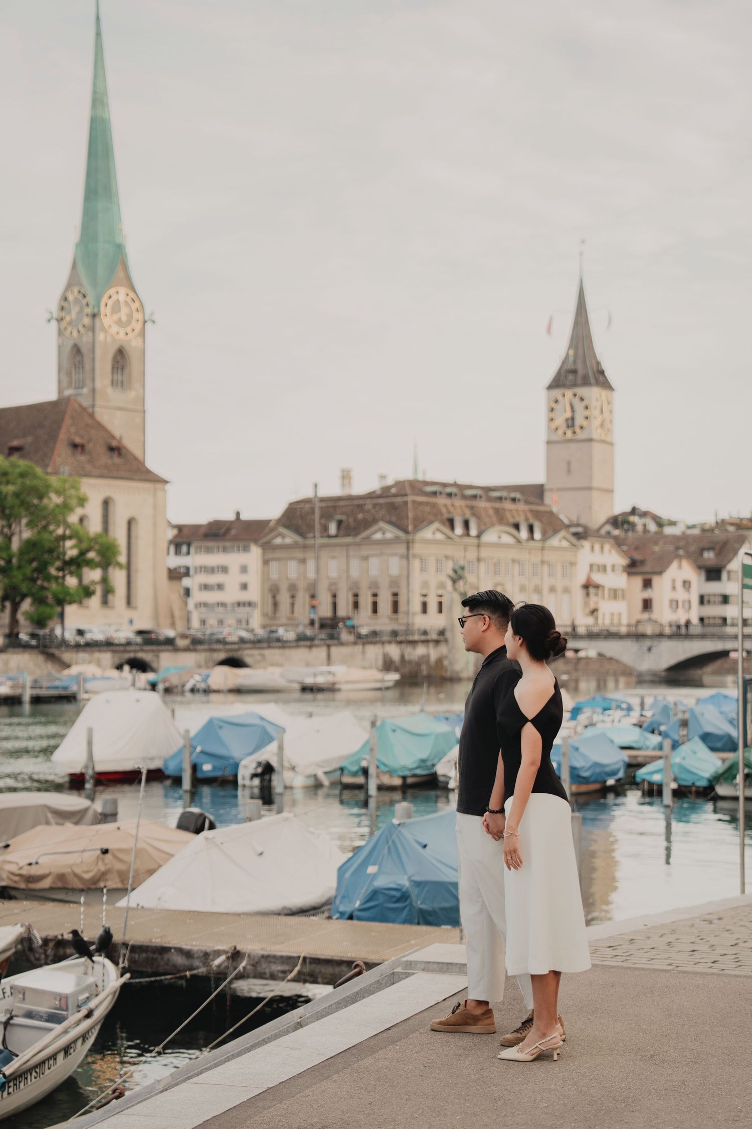 Couple standing by Zurich's boat harbor with Fraumunster and St. Peter's church towers at dusk.