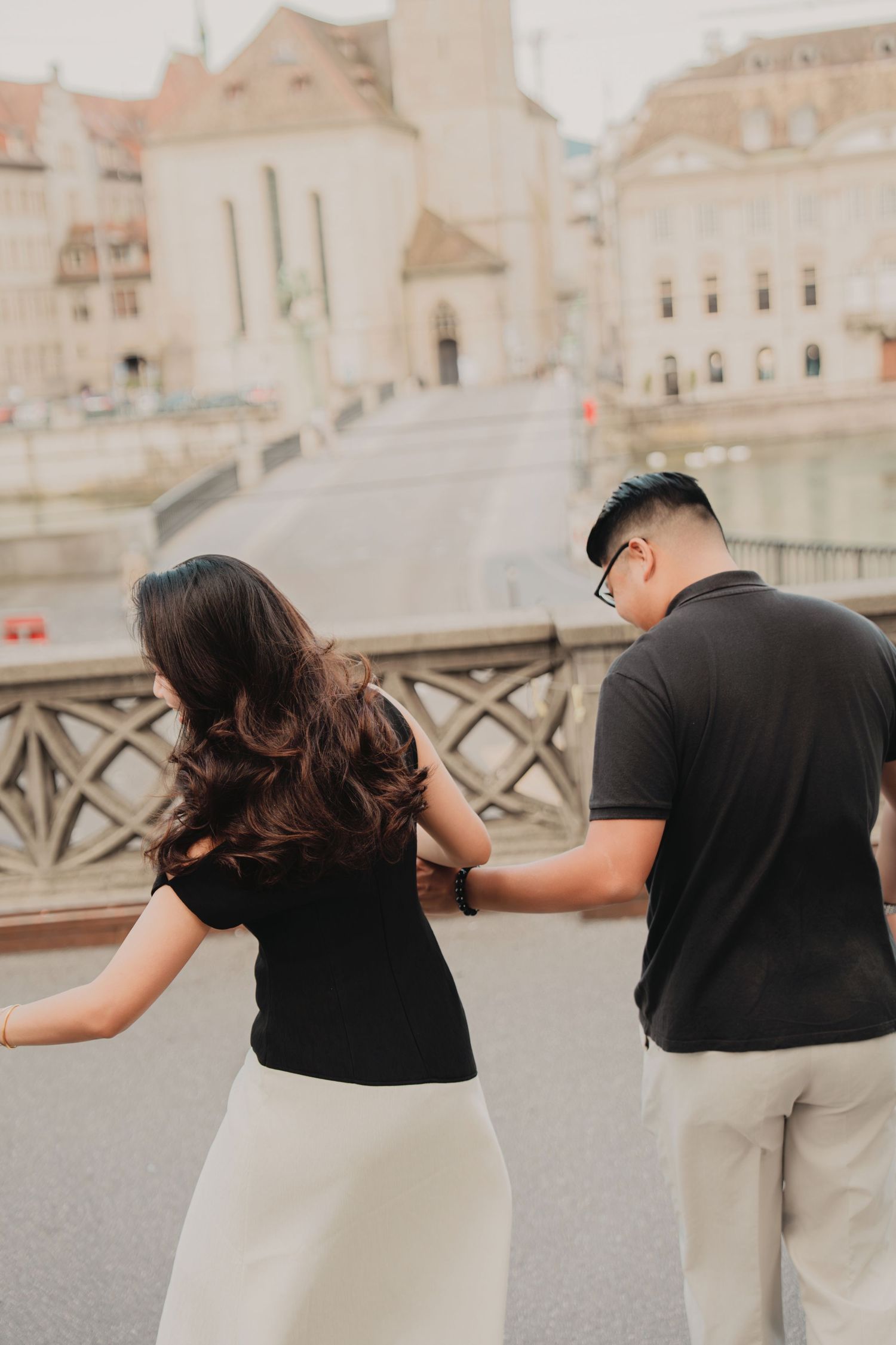 Couple holding hands on a bridge in a European city, woman in black top and white skirt, man in black polo.