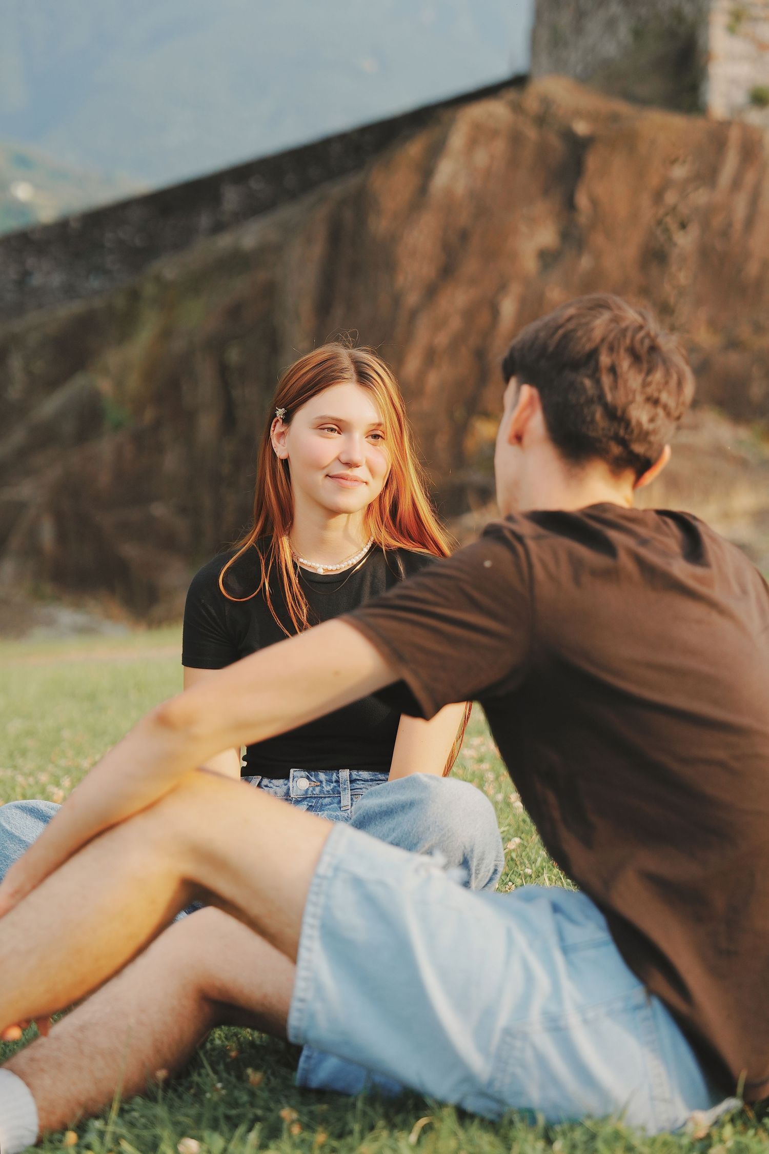 Redhead woman in black tee smiling at partner while sitting together on grass by stone wall.